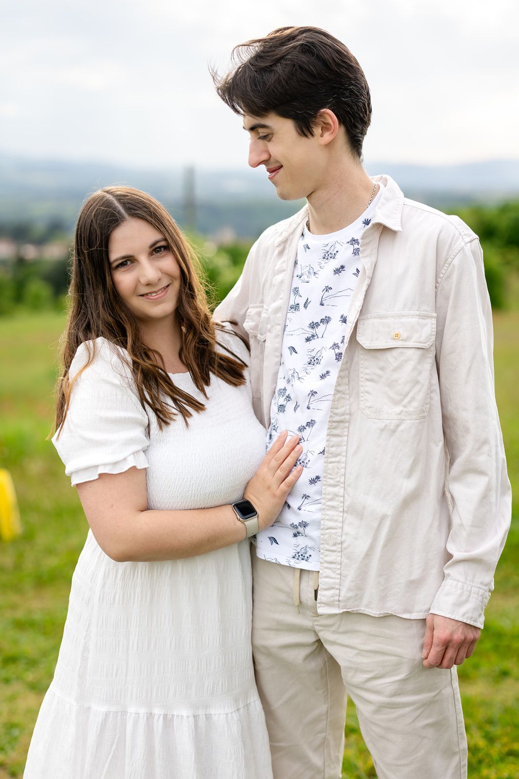 Young couple standing outdoors in a green field, woman in white dress smiling, man in beige shirt looking at her.