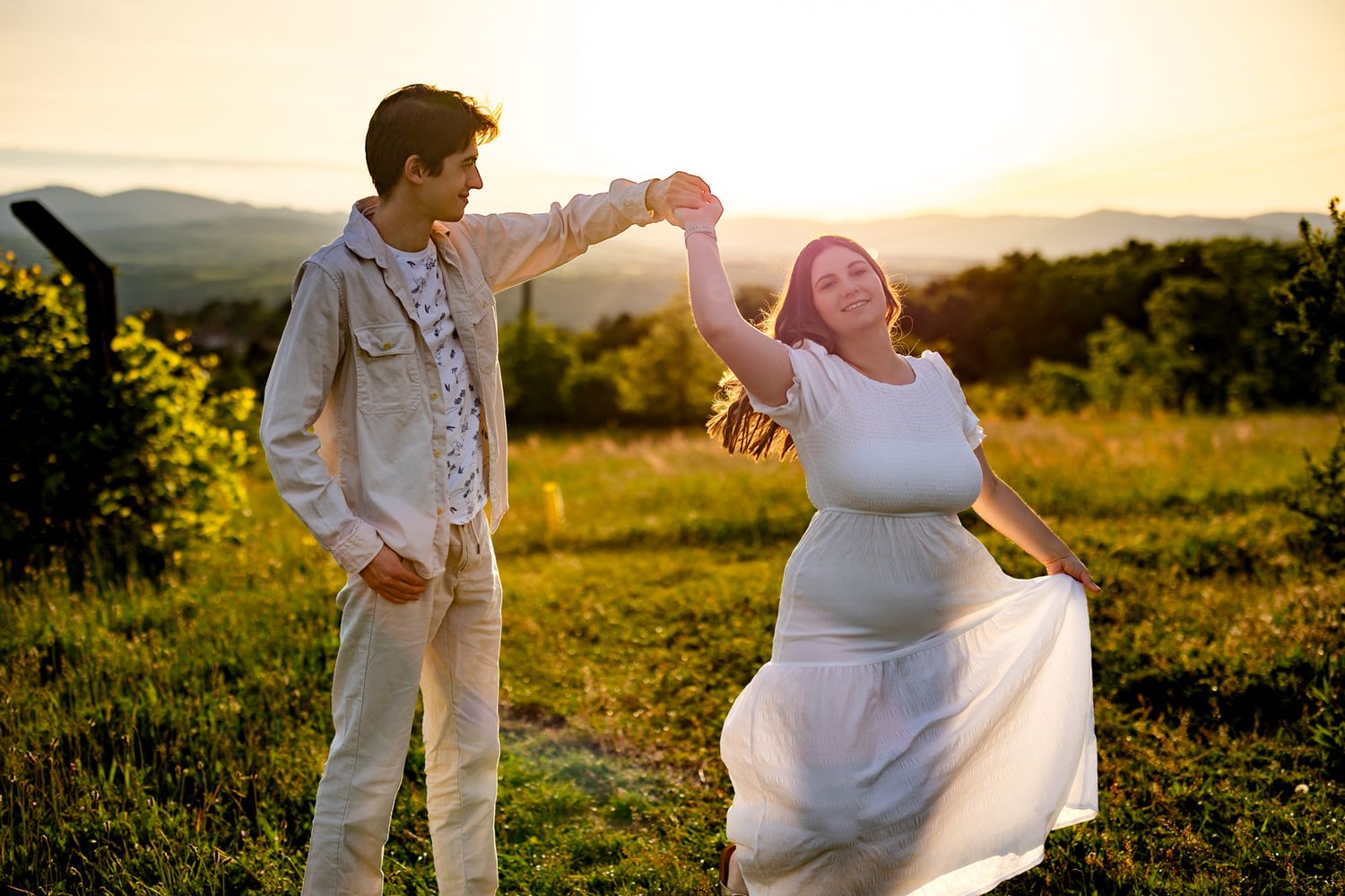 Pregnant woman in white dress dancing with partner at golden sunset in green meadow with mountain views.