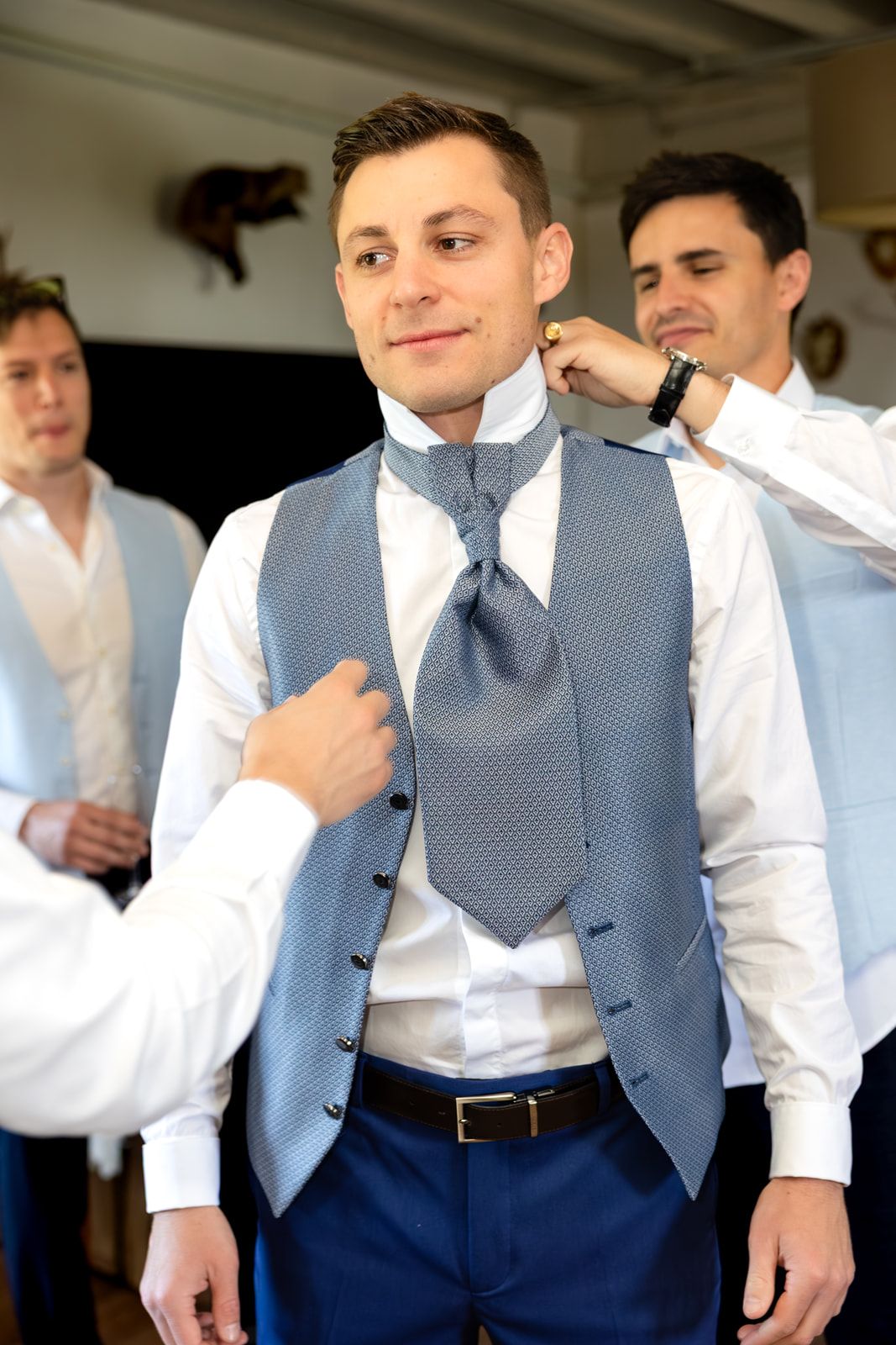 Groomsmen helping groom adjust blue vest and tie before wedding ceremony.