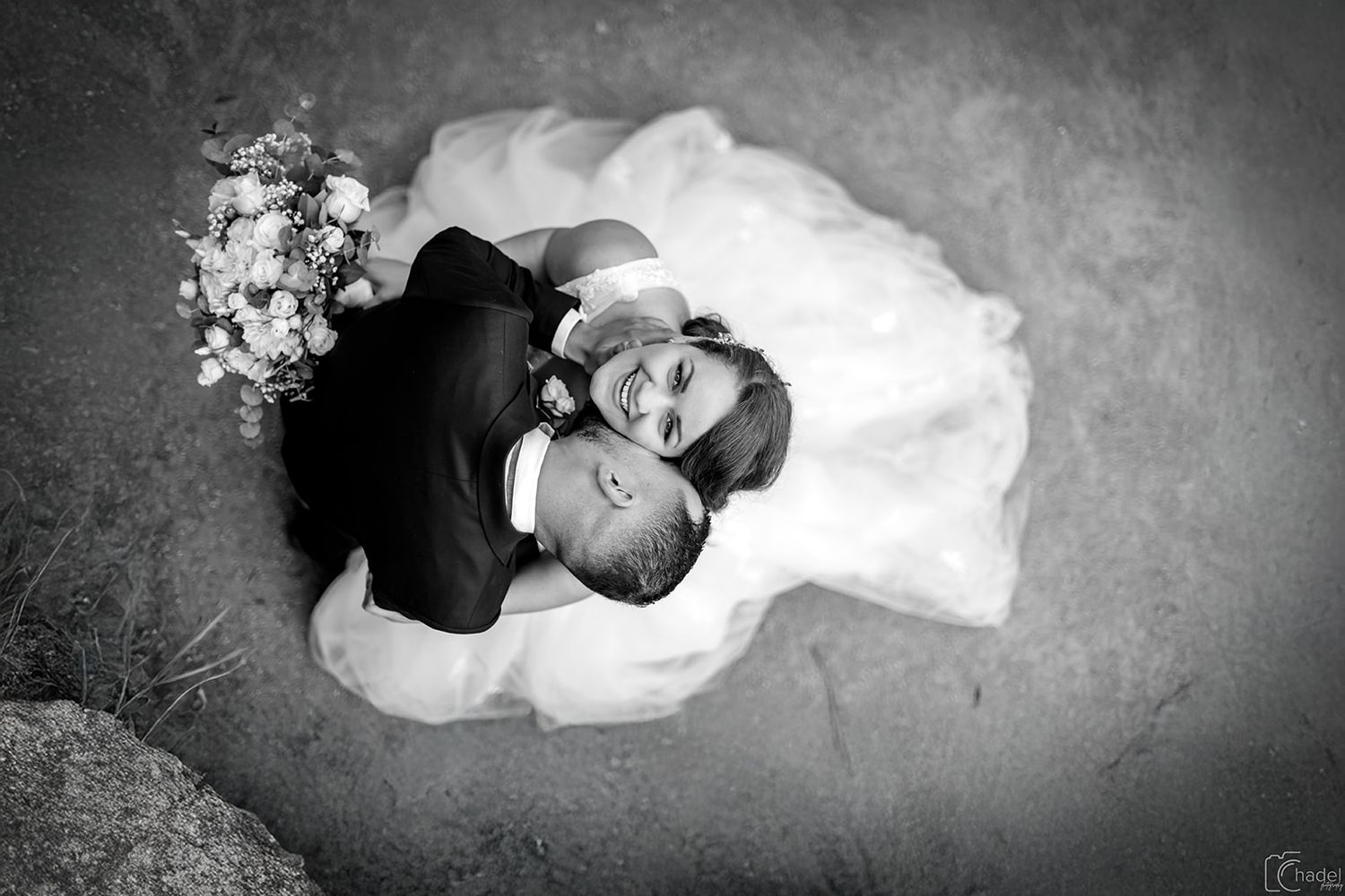 Black and white aerial view of a couple in wedding attire embracing on a textured floor with a bridal bouquet nearby.
