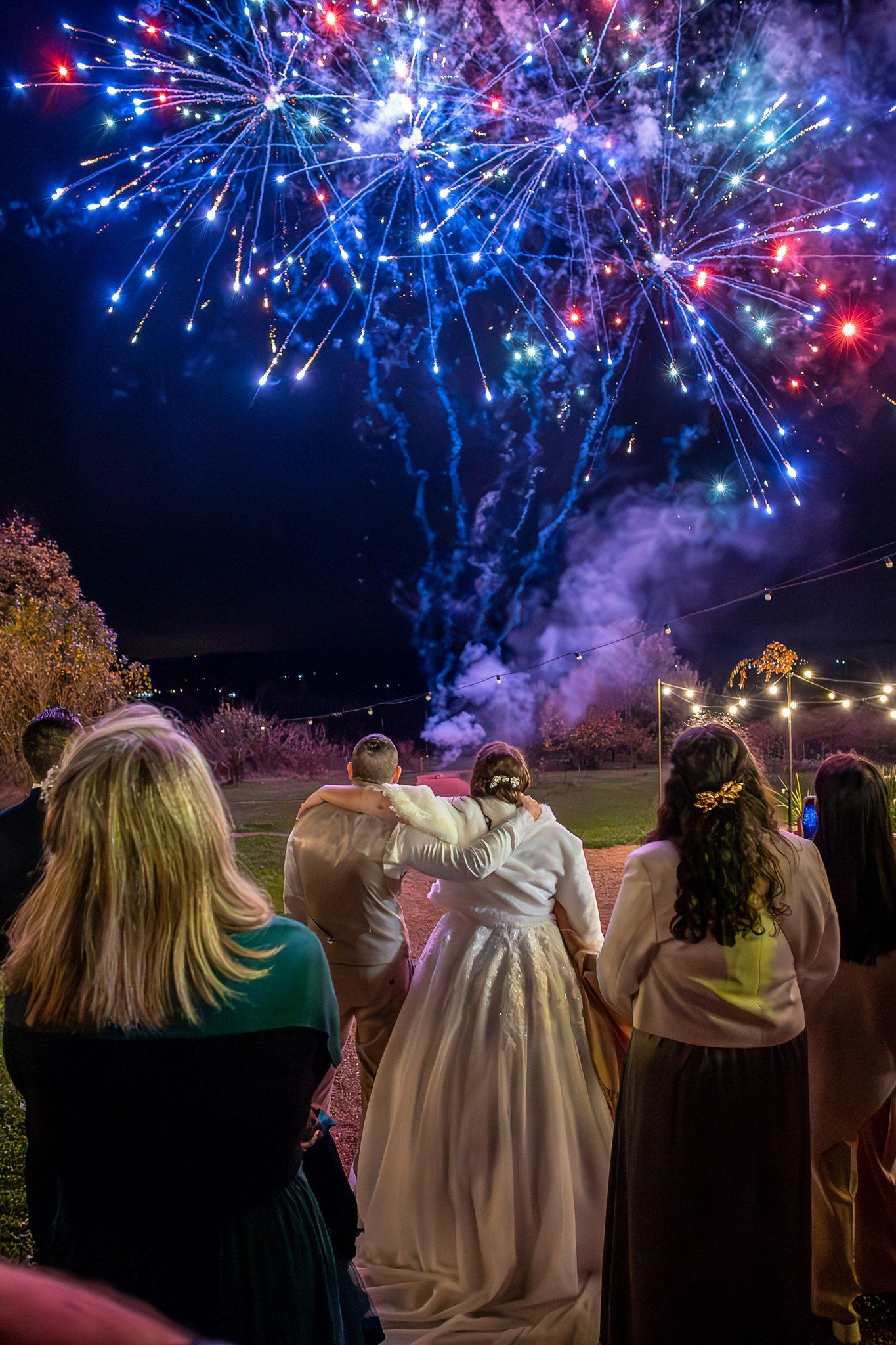 A group of wedding guests watch a spectacular fireworks display with blue and red bursts lighting up the night sky.