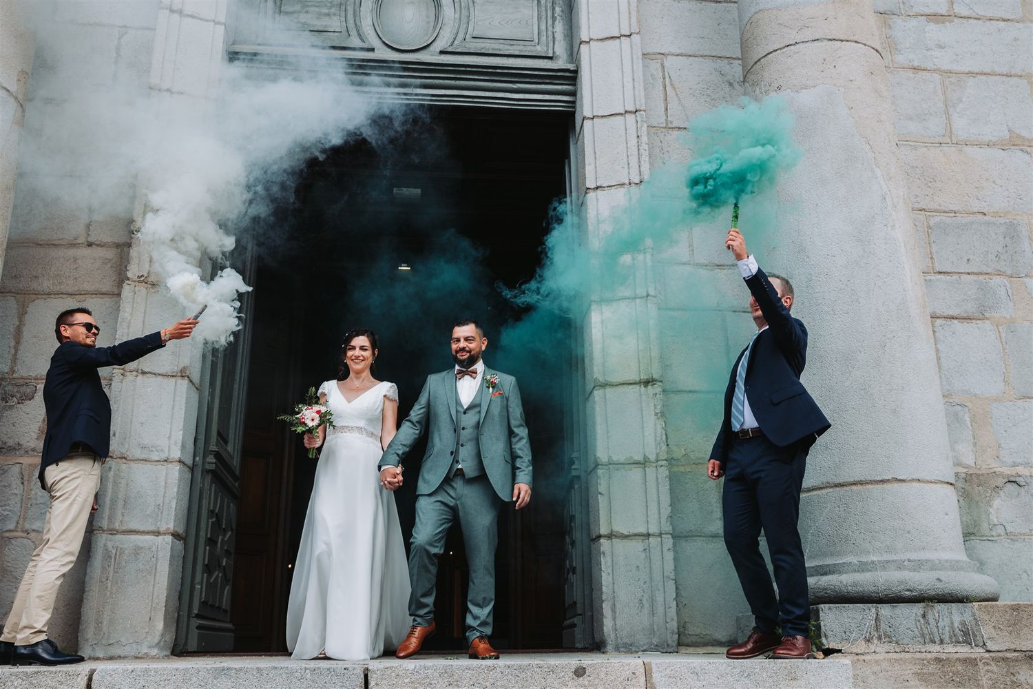 Wedding party poses against stone wall backdrop with colorful smoke bombs creating dramatic teal and orange effects.