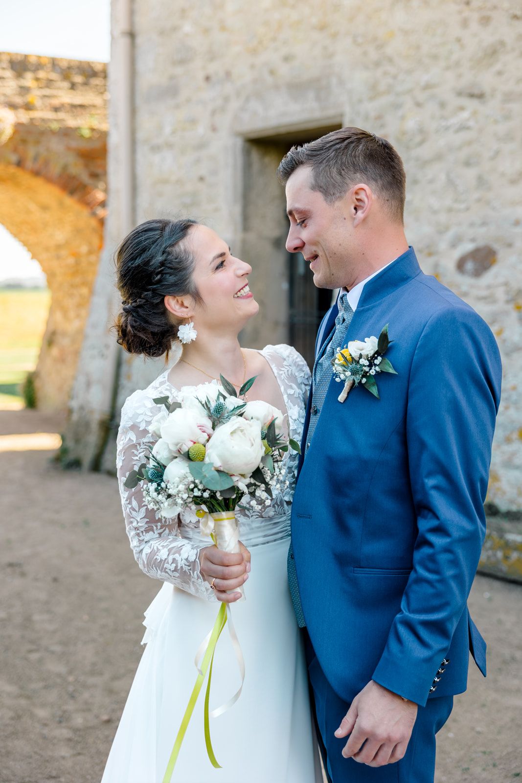 A smiling bride in lace gown holds white peony bouquet while gazing at groom in blue suit near stone arch.