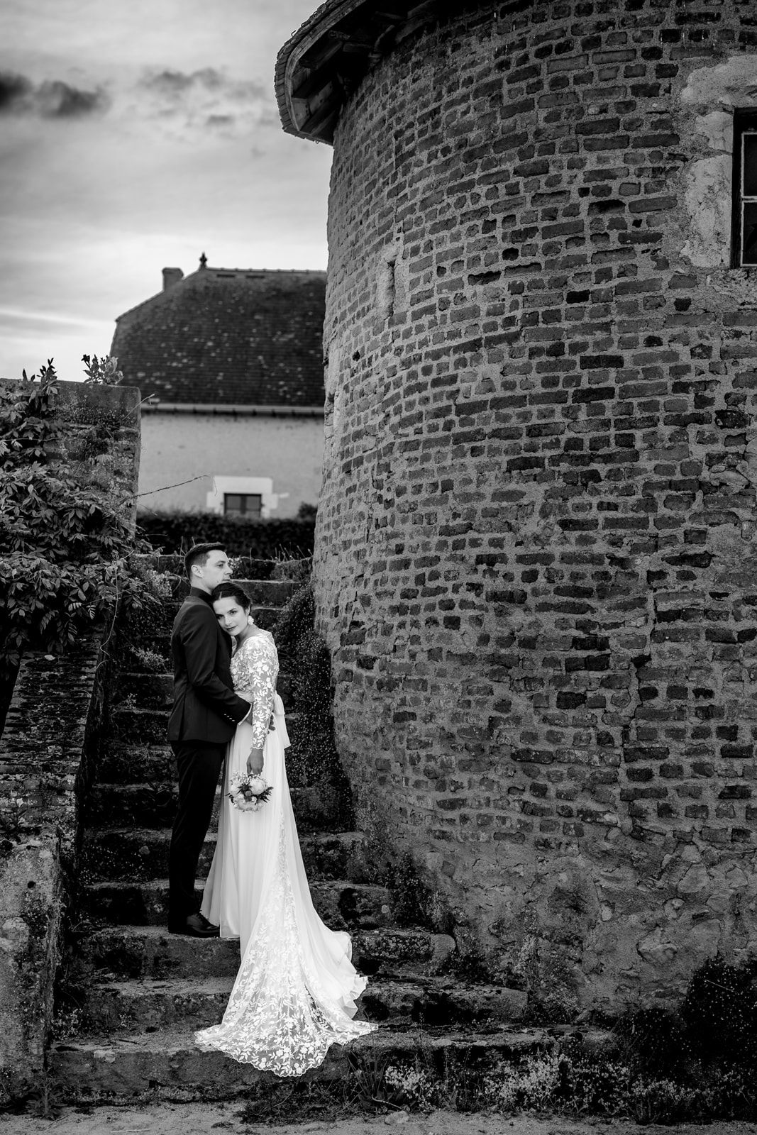 Black and white wedding photo of bride and groom embracing on stone steps beside a historic brick tower.