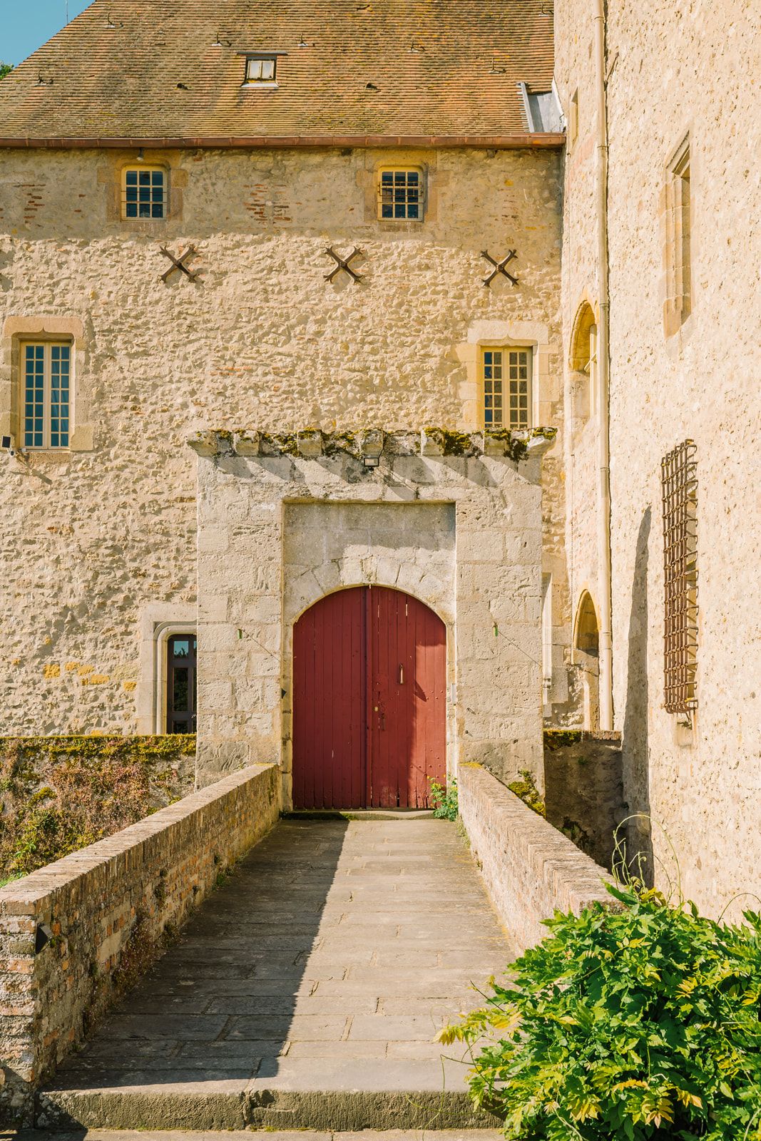 Historic stone castle entrance with red arched door, stone pathway, and lush greenery under warm sunlight.