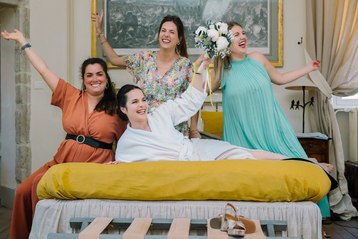 Joyful bride and bridesmaids celebrating on a bed, holding bouquet and cheering together.