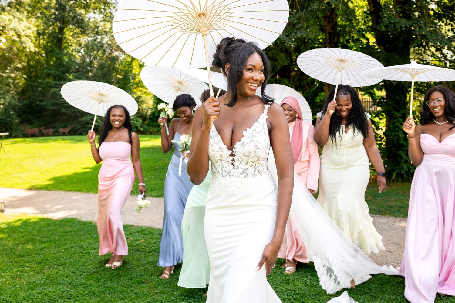 Wedding party poses with white parasols on sunny lawn wearing elegant dresses in pink, blue and white tones.