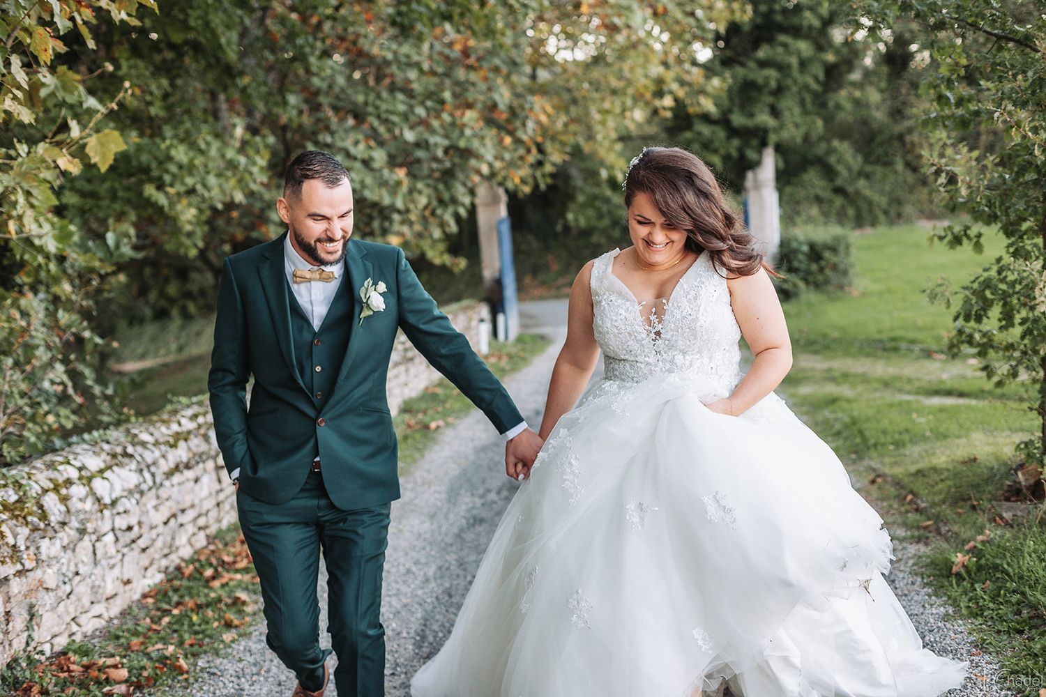 A couple in wedding attire walks through a scenic outdoor path surrounded by greenery and natural light.