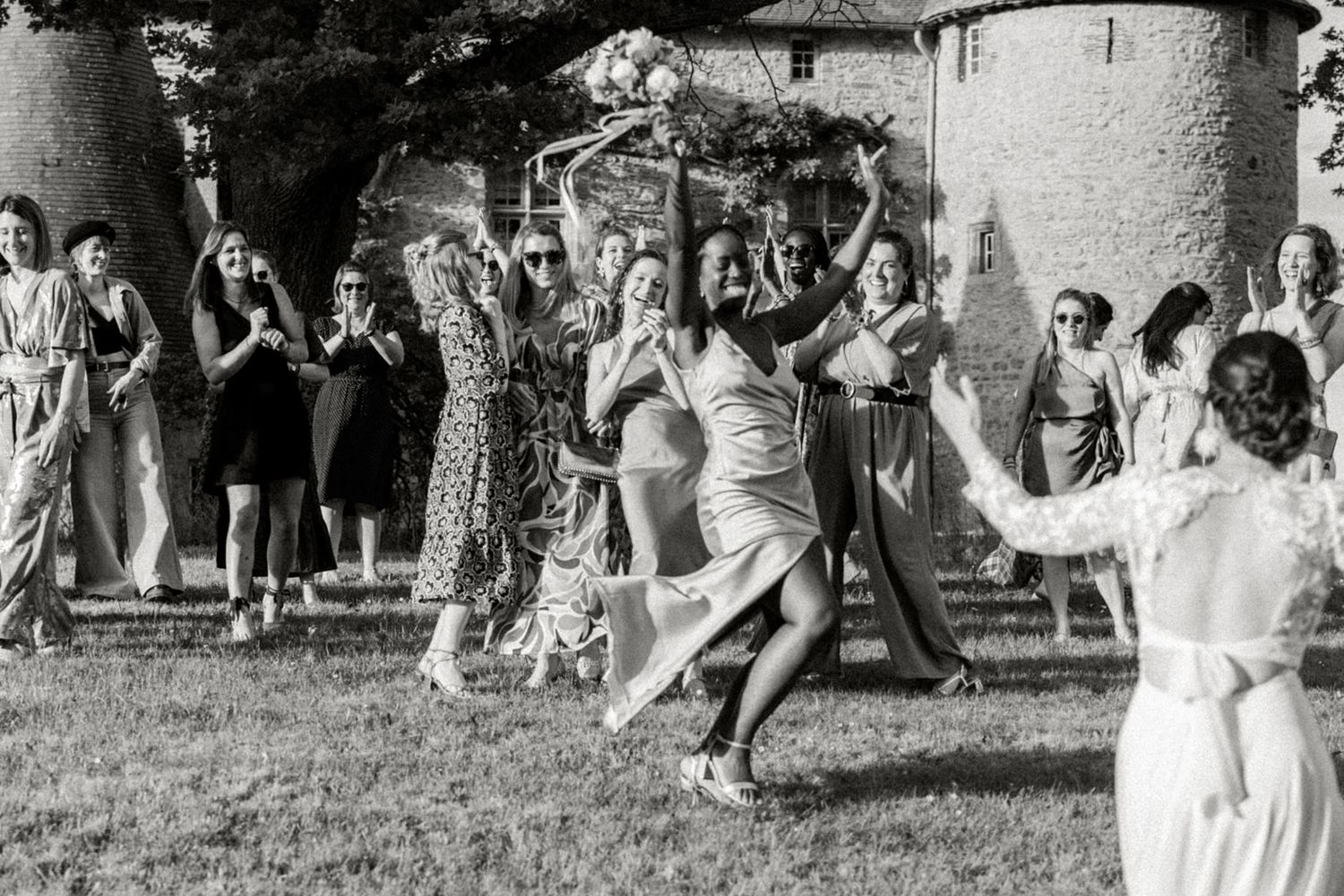 Black and white photo of wedding guests laughing as bride tosses bouquet outdoors near stone building.