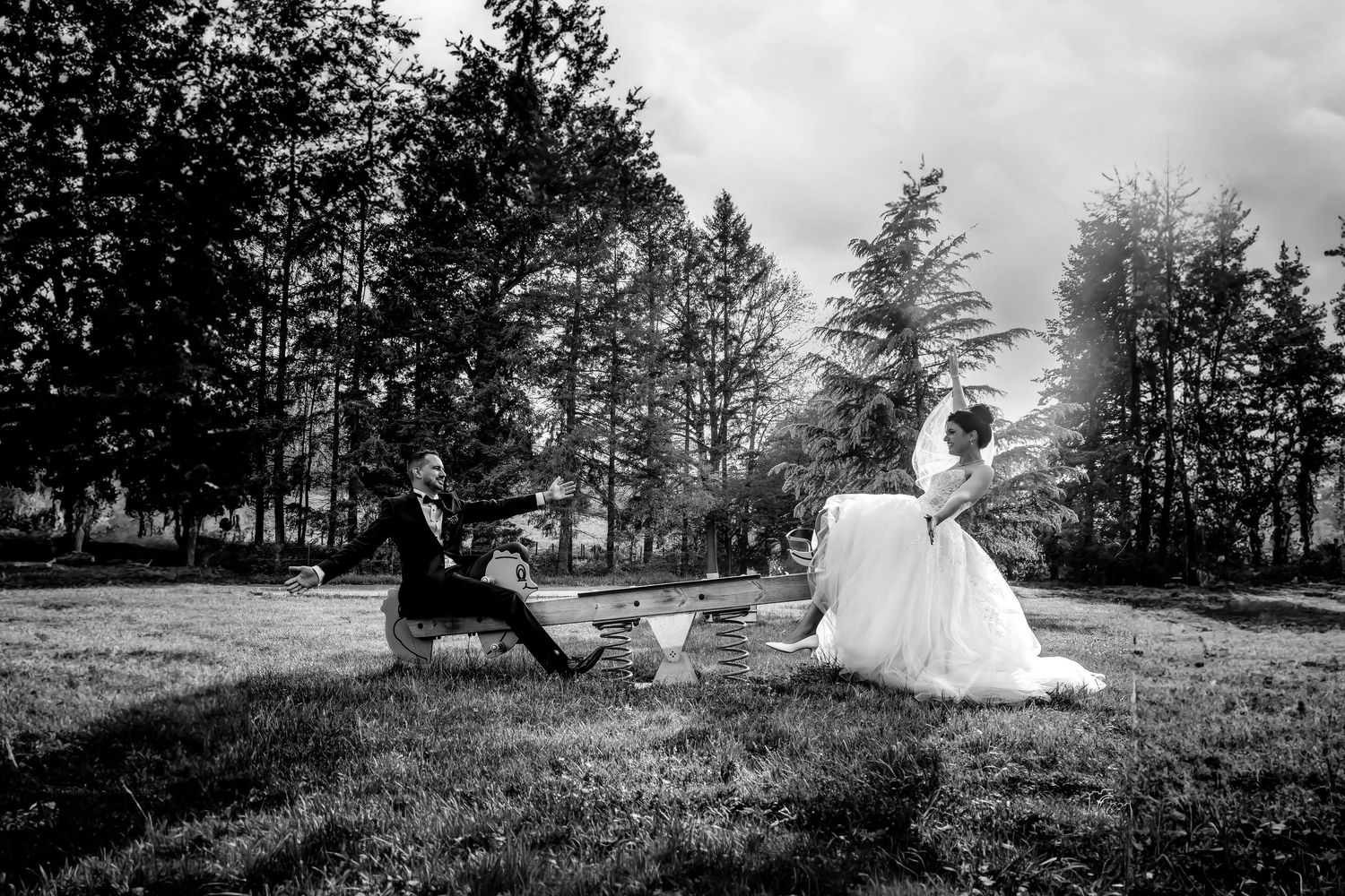 A black and white wedding photo shows a playful moment between a couple in a grassy field surrounded by tall trees.