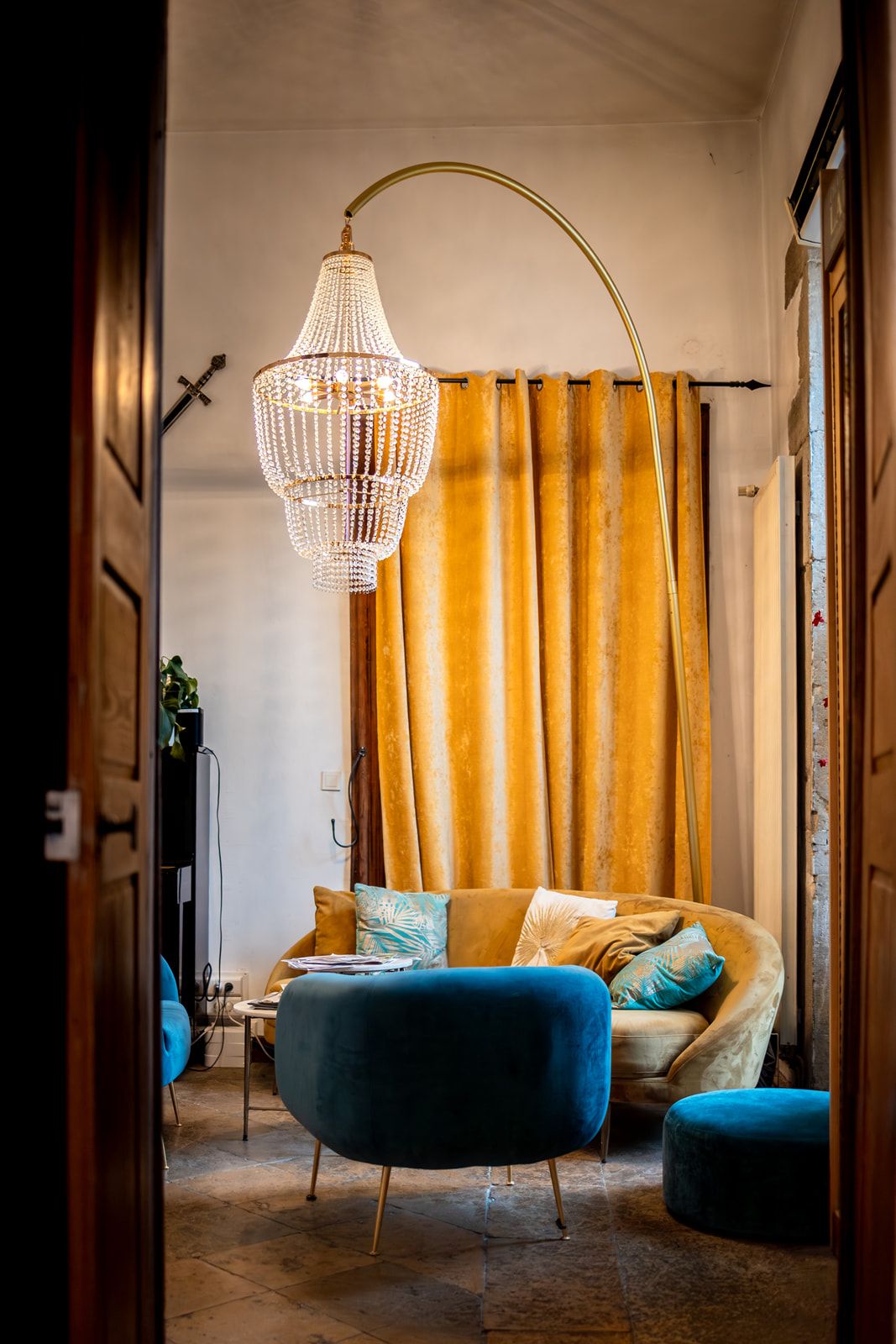 Elegant living room with curved velvet sofa, crystal chandelier, and mustard yellow curtains against white walls.