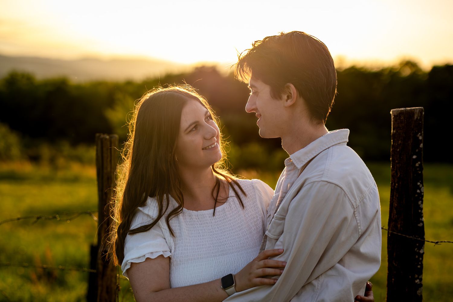 Couple embracing at golden sunset in a green field, gazing lovingly at each other near a rustic fence.