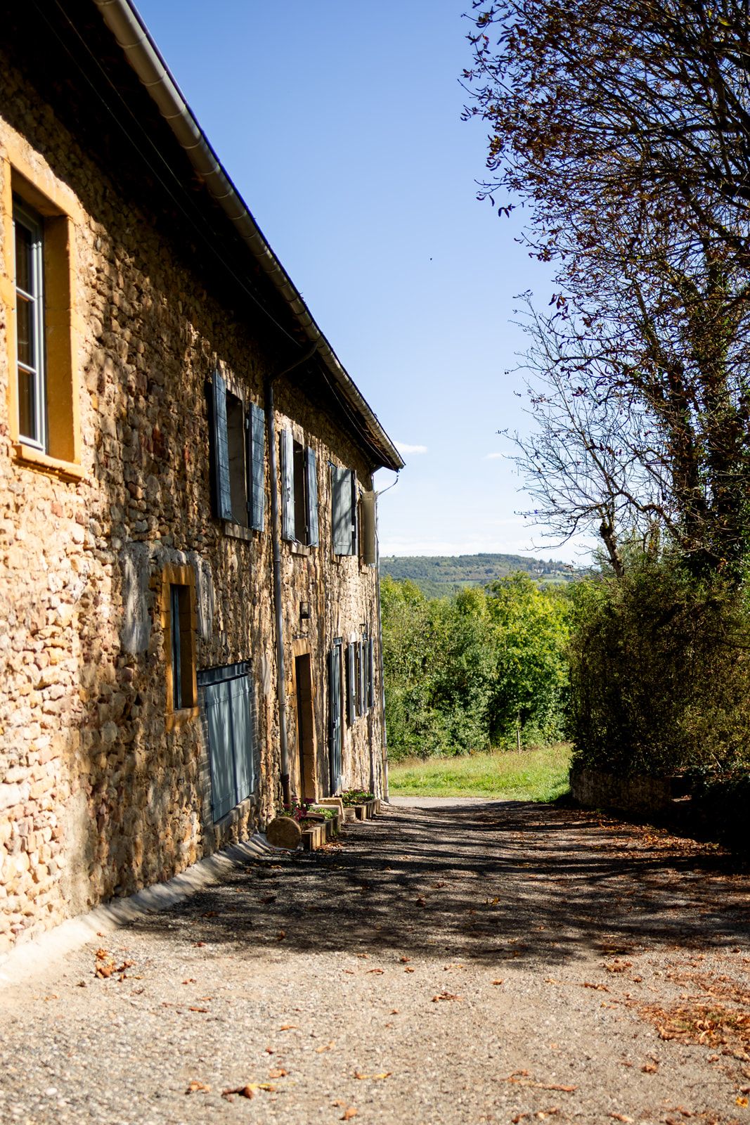 Long rustic stone building with blue shuttered windows and gravel path surrounded by countryside scenery.
