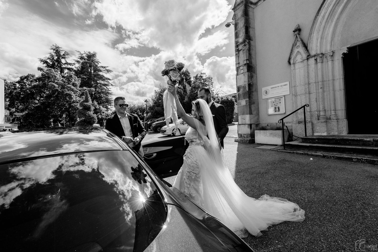Black and white wedding photo showing a couple exiting a vintage car outside a church on a sunny day.