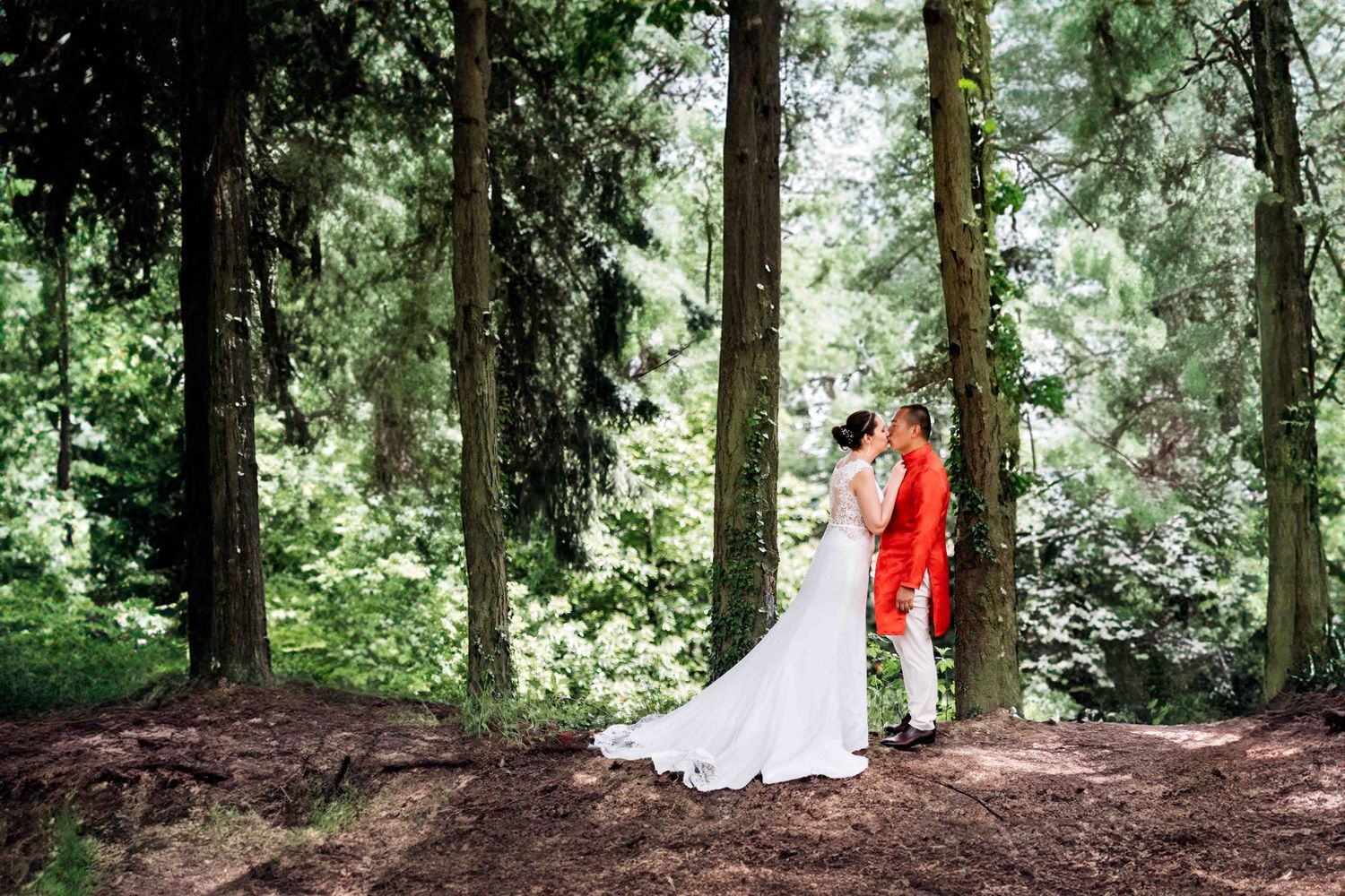 A scenic forest wedding photoshoot with a white dress and red accent visible through tall evergreen trees.