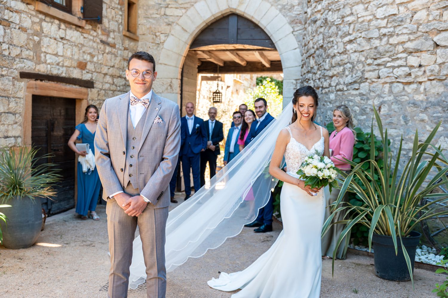 A wedding party poses under a stone archway at a rustic venue with decorative plants and flowing white veil.