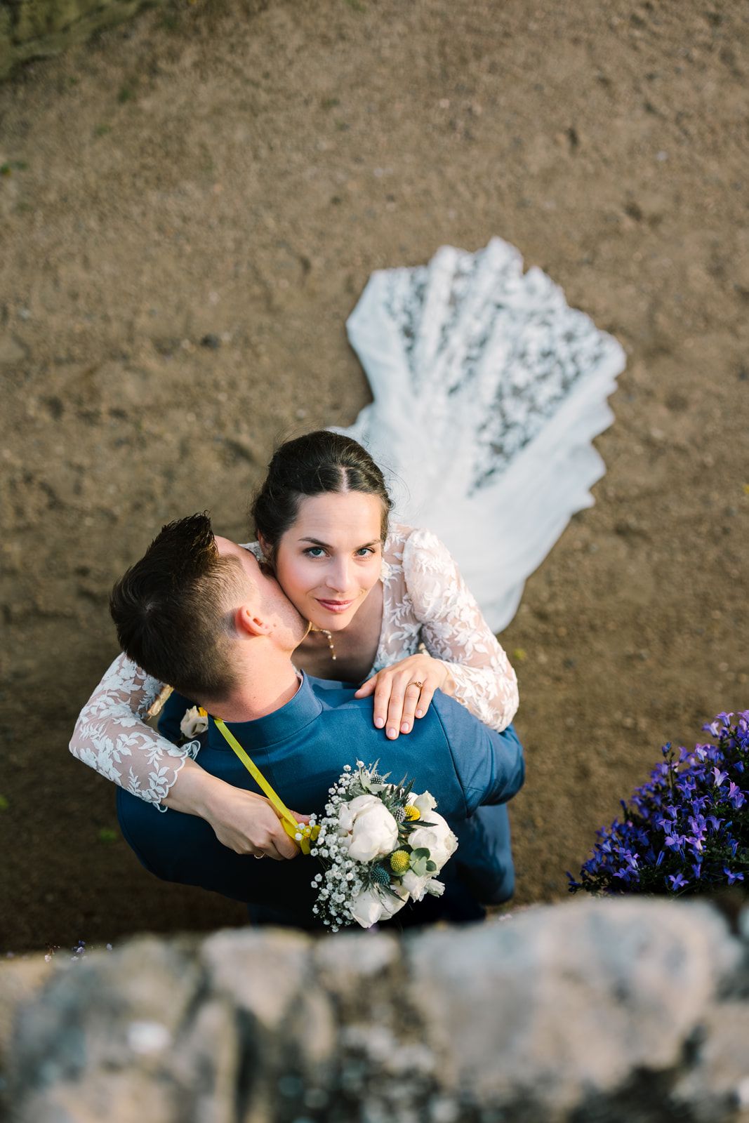 Aerial view of newlyweds embracing outdoors, bride in lace dress with veil spread, holding white bouquet near purple flowers.