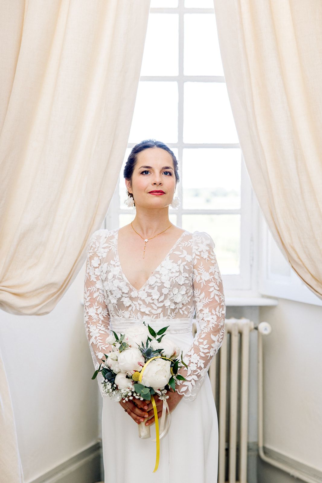 Elegant bride in lace long-sleeve wedding dress holding white floral bouquet standing by a sunlit window.