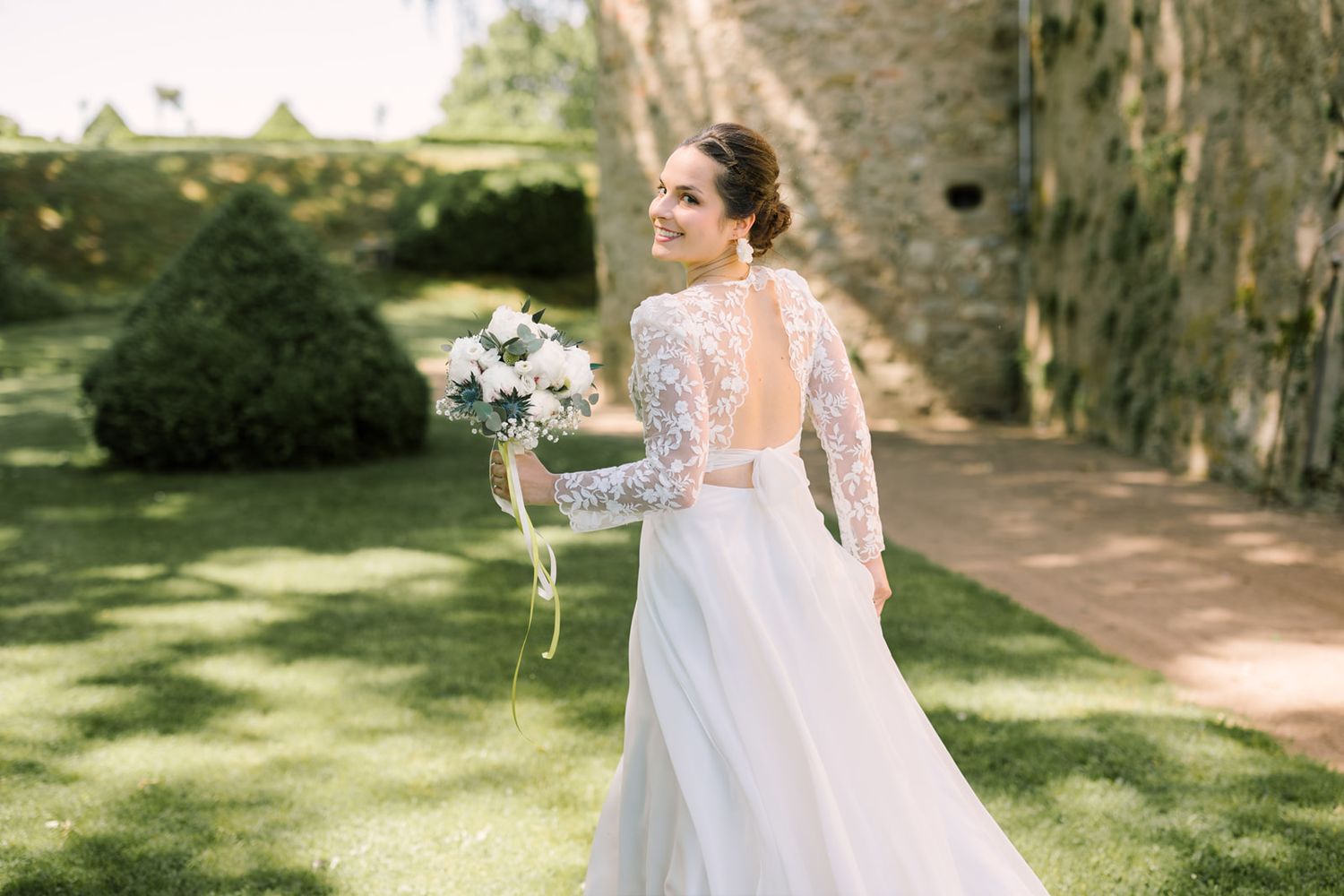 Smiling bride in lace long-sleeve wedding gown holding white bouquet, turning gracefully in a lush garden setting.