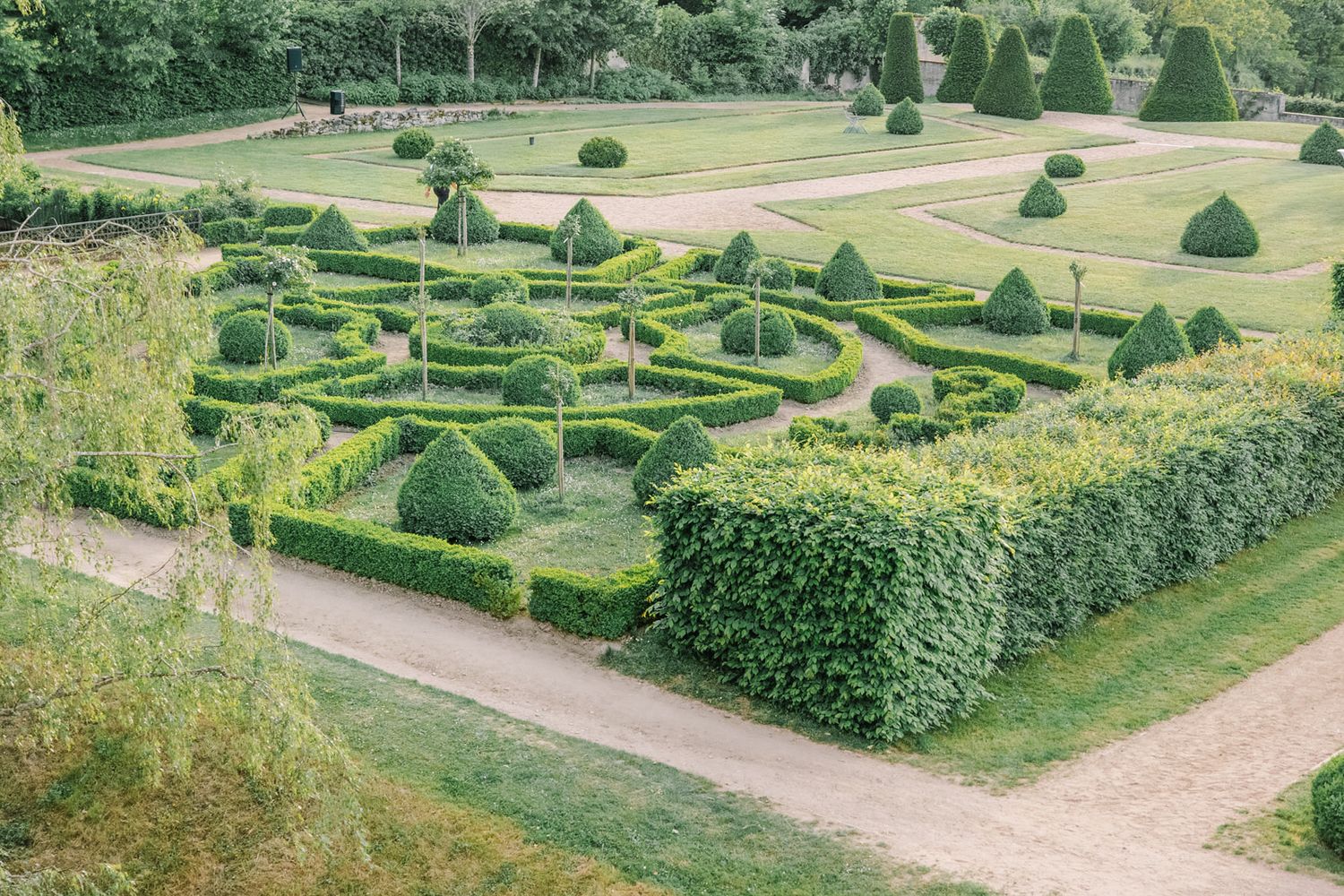Aerial view of a formal French garden with geometric topiary hedges, cone-shaped shrubs, and manicured green lawns.