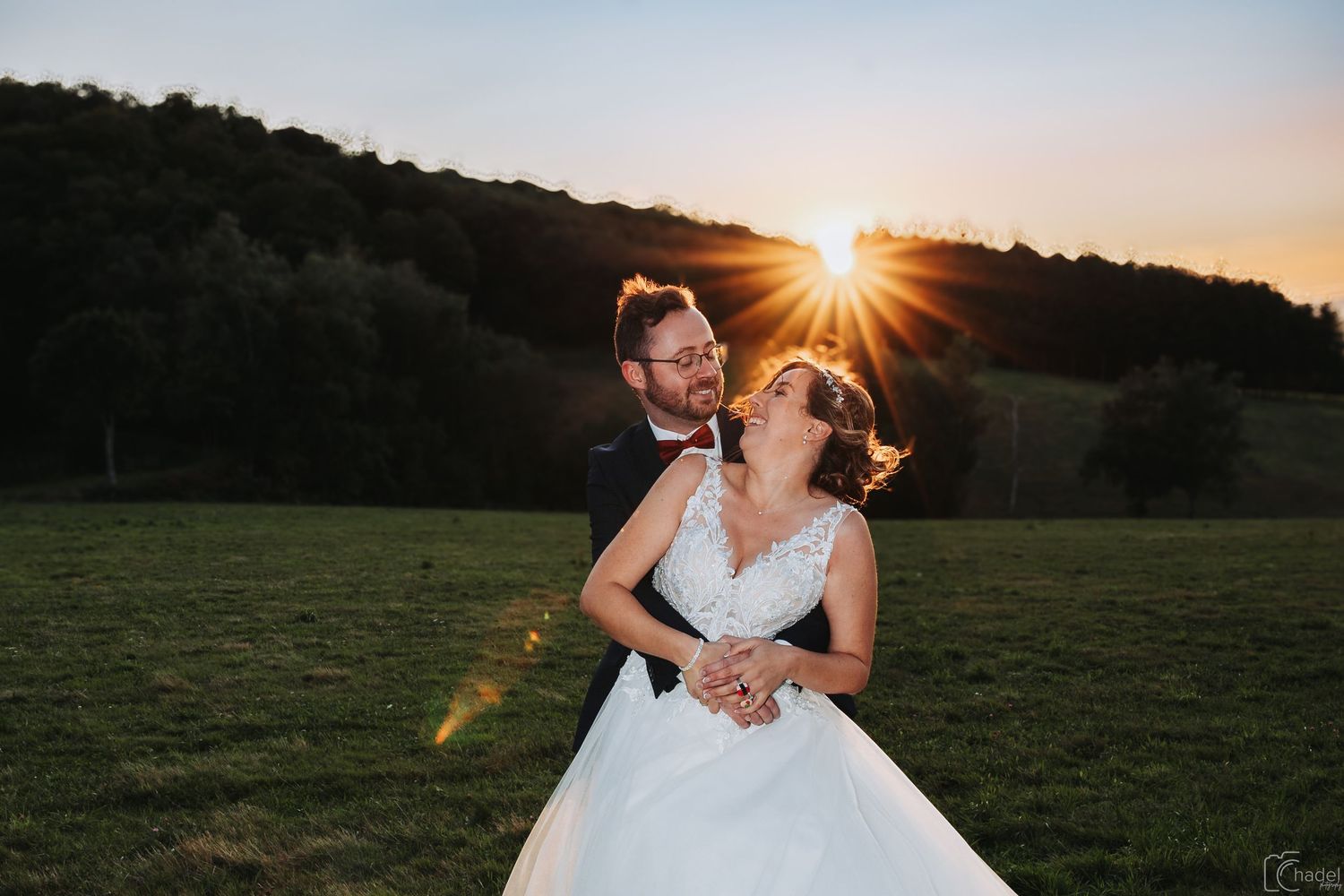 A couple in wedding attire dances together in a sunlit meadow at golden hour.