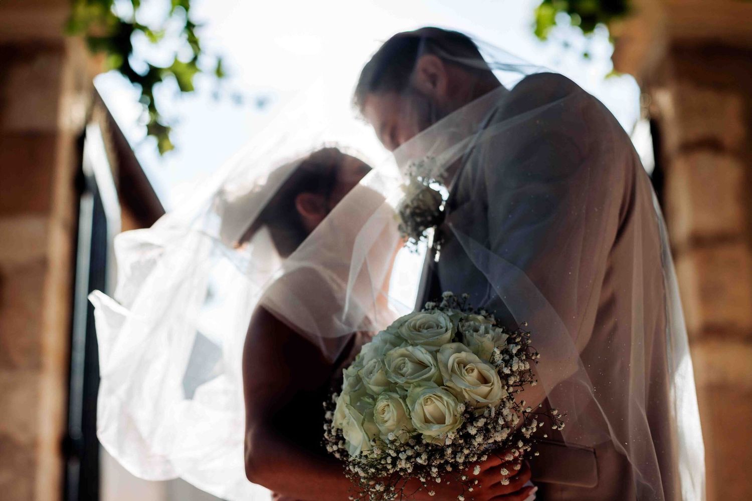 A tender wedding moment captured under sunlit trees with a flowing white veil.