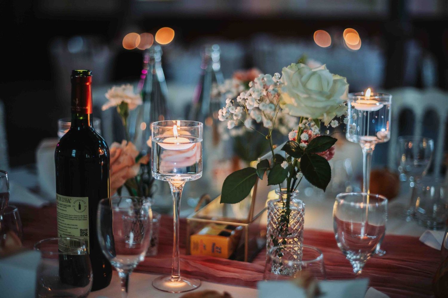 Romantic table setting with wine bottle, glowing candles, and delicate white flowers in a glass vase at dusk.