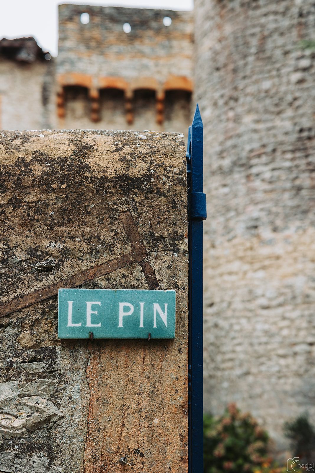 A rustic wooden post with a weathered blue sign reading 'LE PIN' mounted against an old stone wall background.