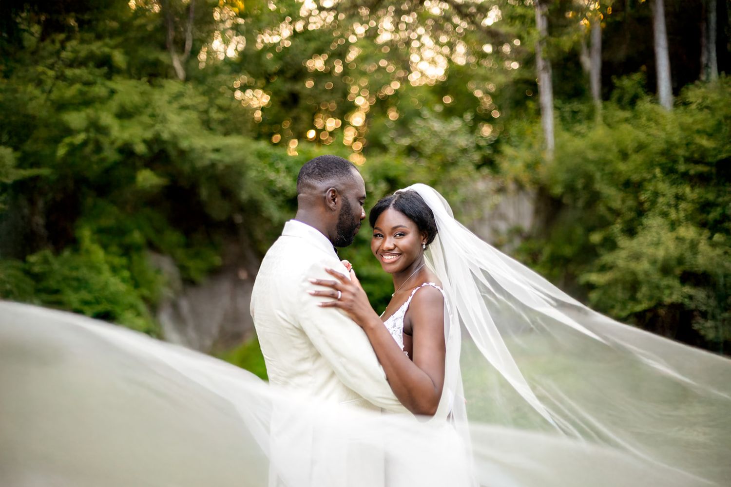 A romantic wedding couple embraces in a lush garden setting as a white veil flows elegantly in the breeze.
