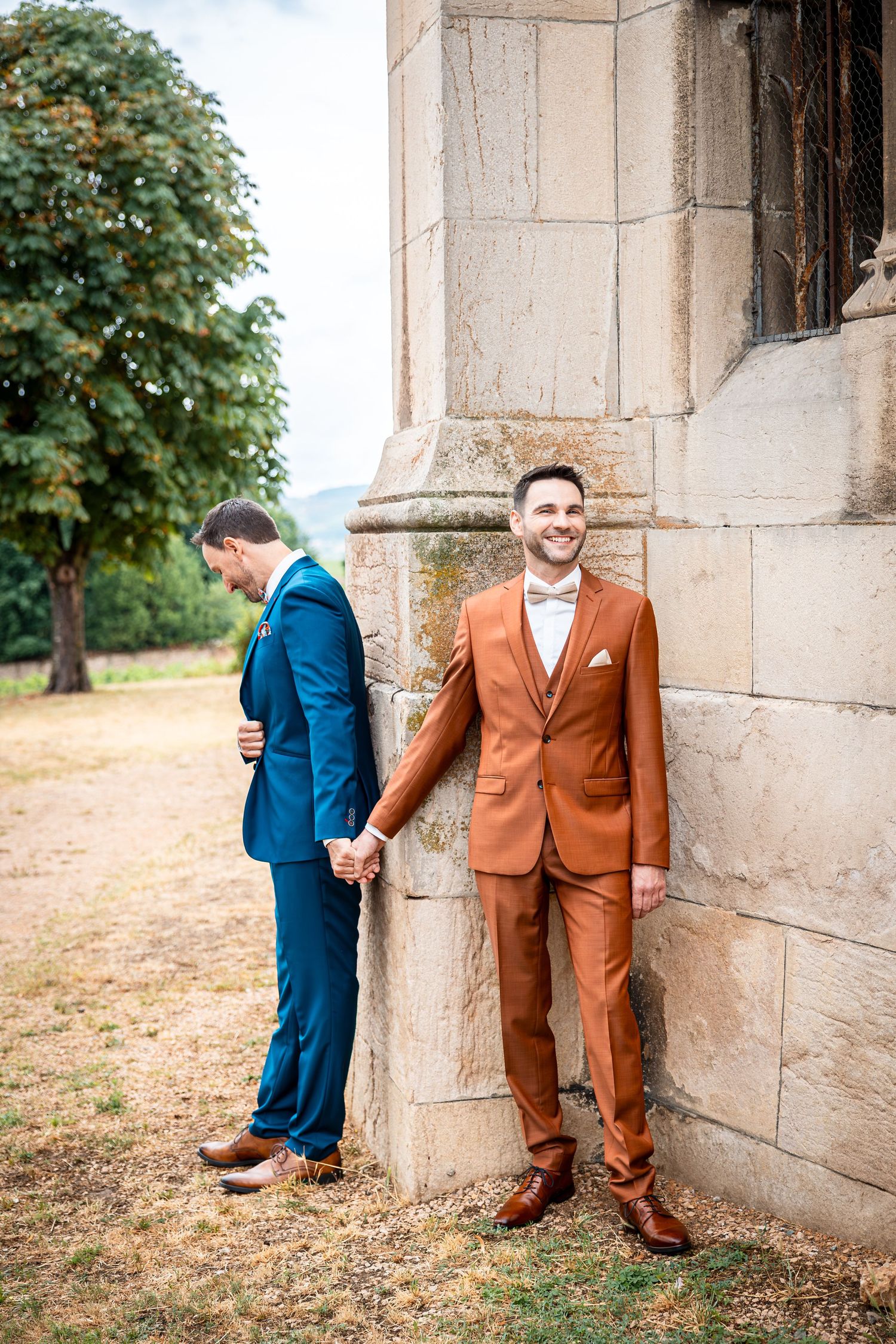 Two grooms in contrasting navy and rust-colored suits pose playfully on opposite sides of a stone pillar outdoors.