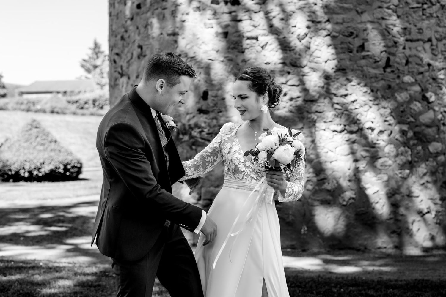 Black and white photo of a bride holding a bouquet and groom holding hands outdoors near a stone wall.