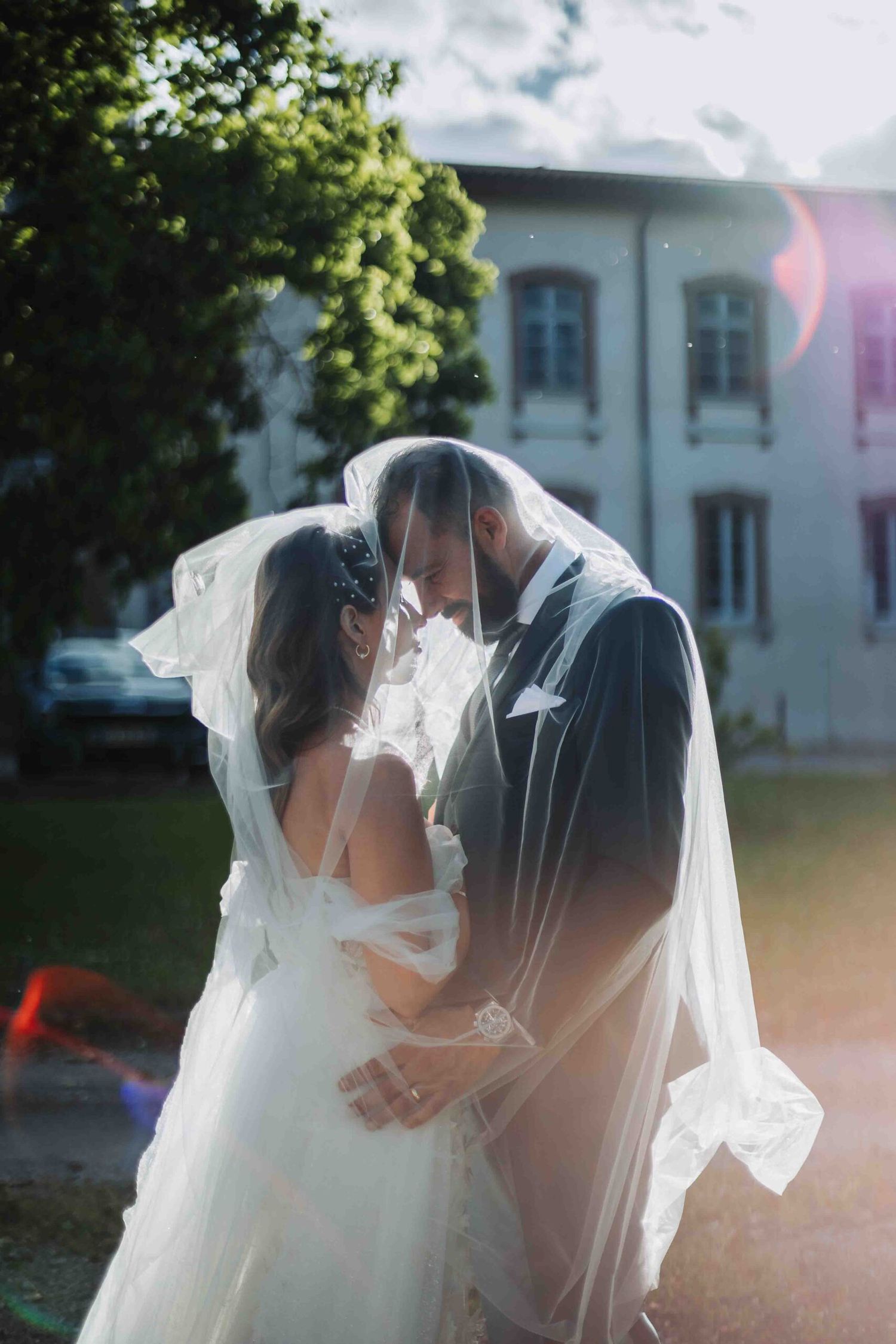 Romantic wedding couple embracing under flowing veil in garden setting.
