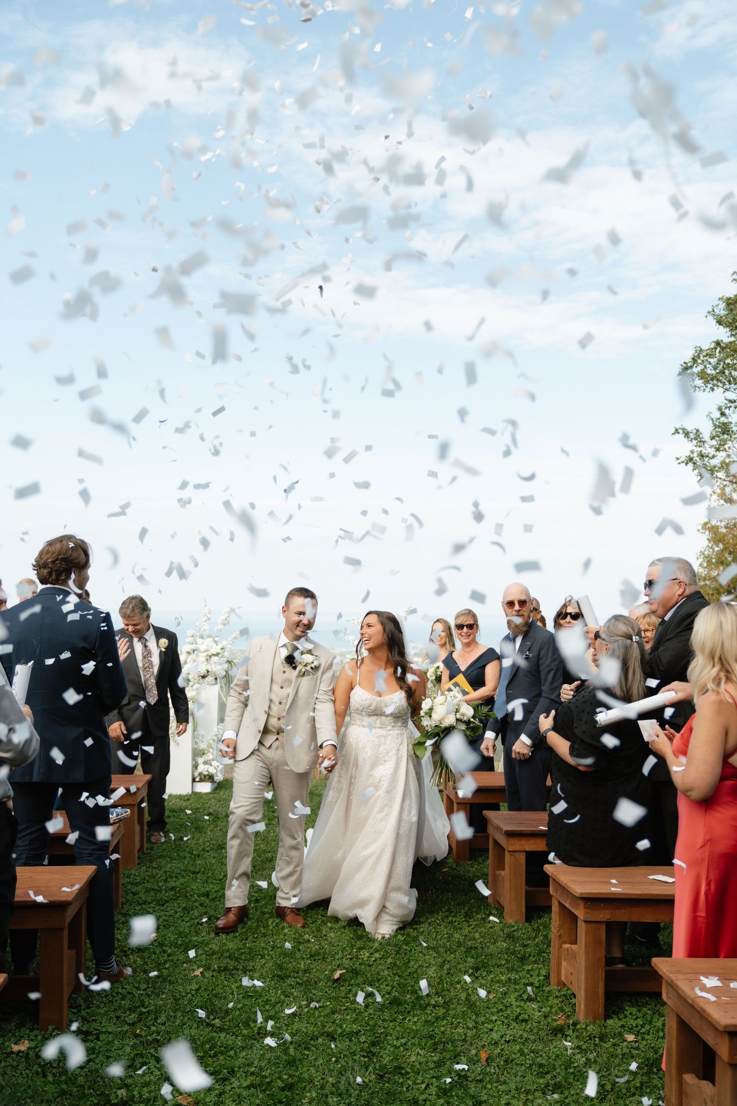 Wedding guests shower the newlyweds with white confetti during an outdoor ceremony.