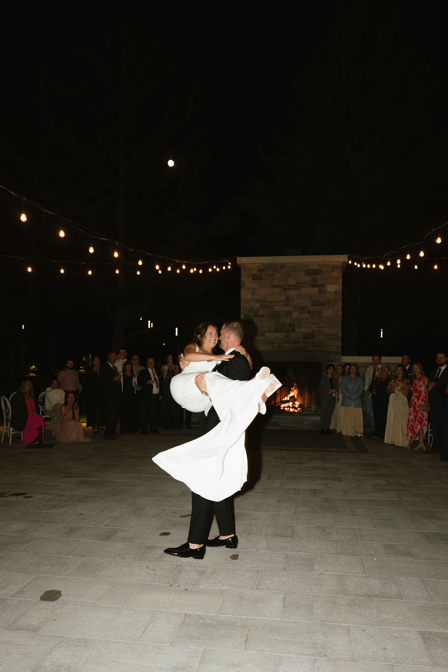 A couple shares their first dance under string lights at an outdoor evening wedding reception.