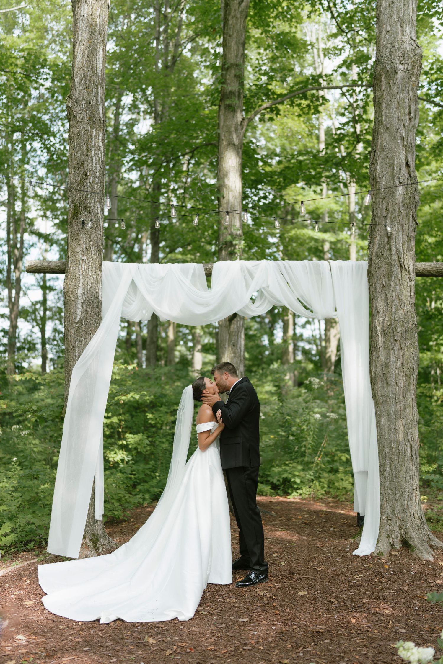 Bride and groom share first kiss under white draped arch surrounded by lush green trees in an outdoor forest wedding ceremony.