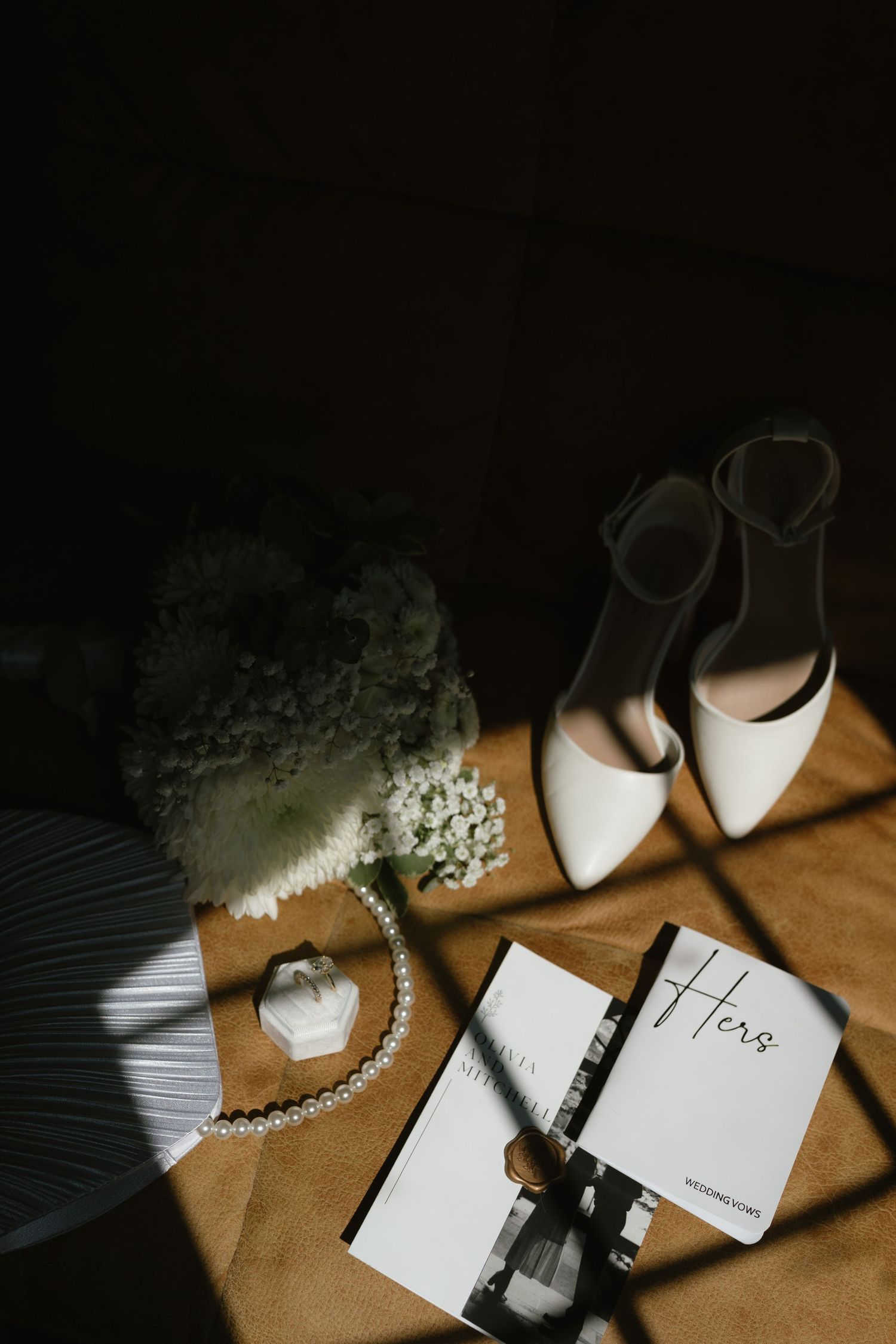 Bridal flat lay with white heels, pearl necklace, wedding bouquet, ring box, and Hers vow book on golden surface.