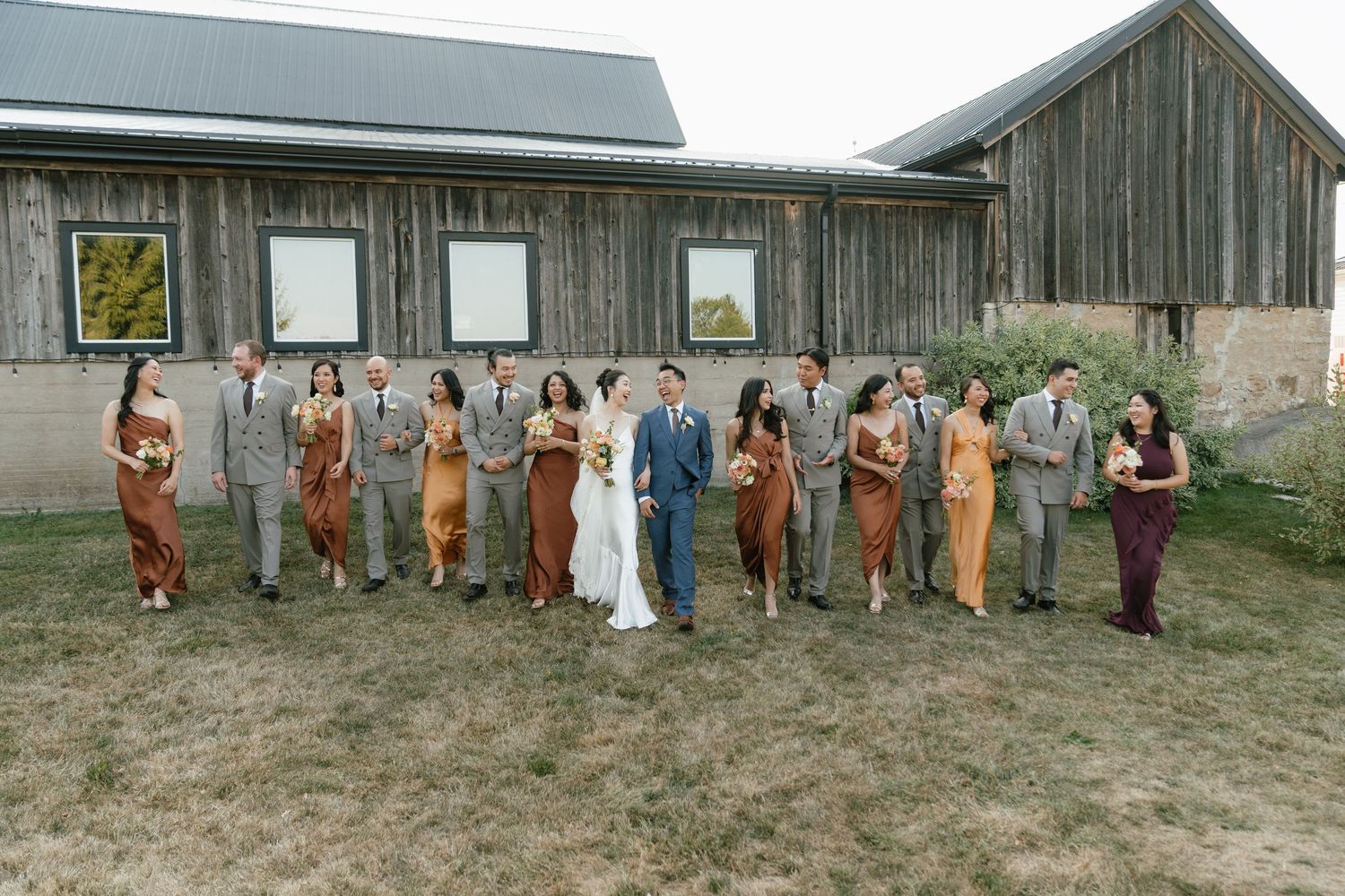 Wedding party poses in front of a rustic barn wearing coordinated outfits.