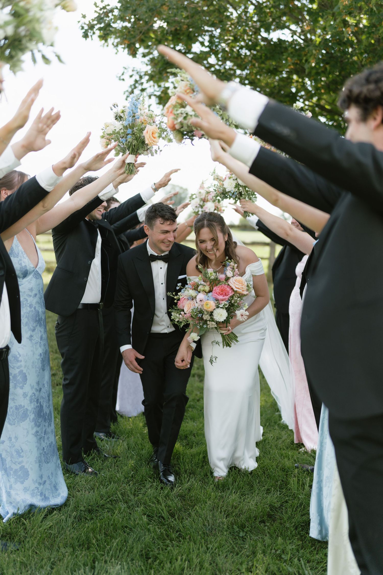 Bride and groom walk through an arch of raised hands formed by wedding party on green grass.