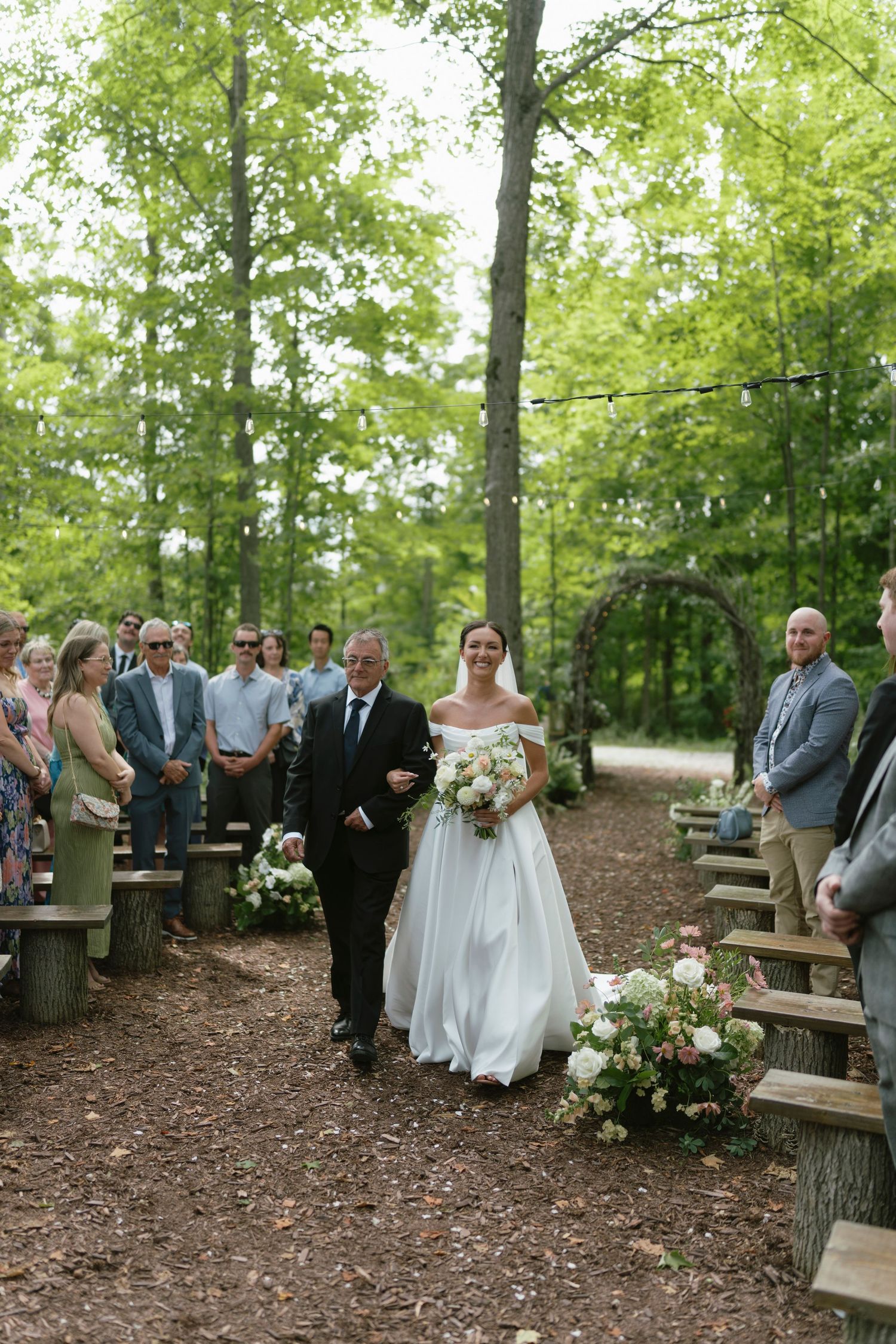 Bride walks down wooded outdoor aisle with escort, holding bouquet, guests seated on log benches, floral arch in background.
