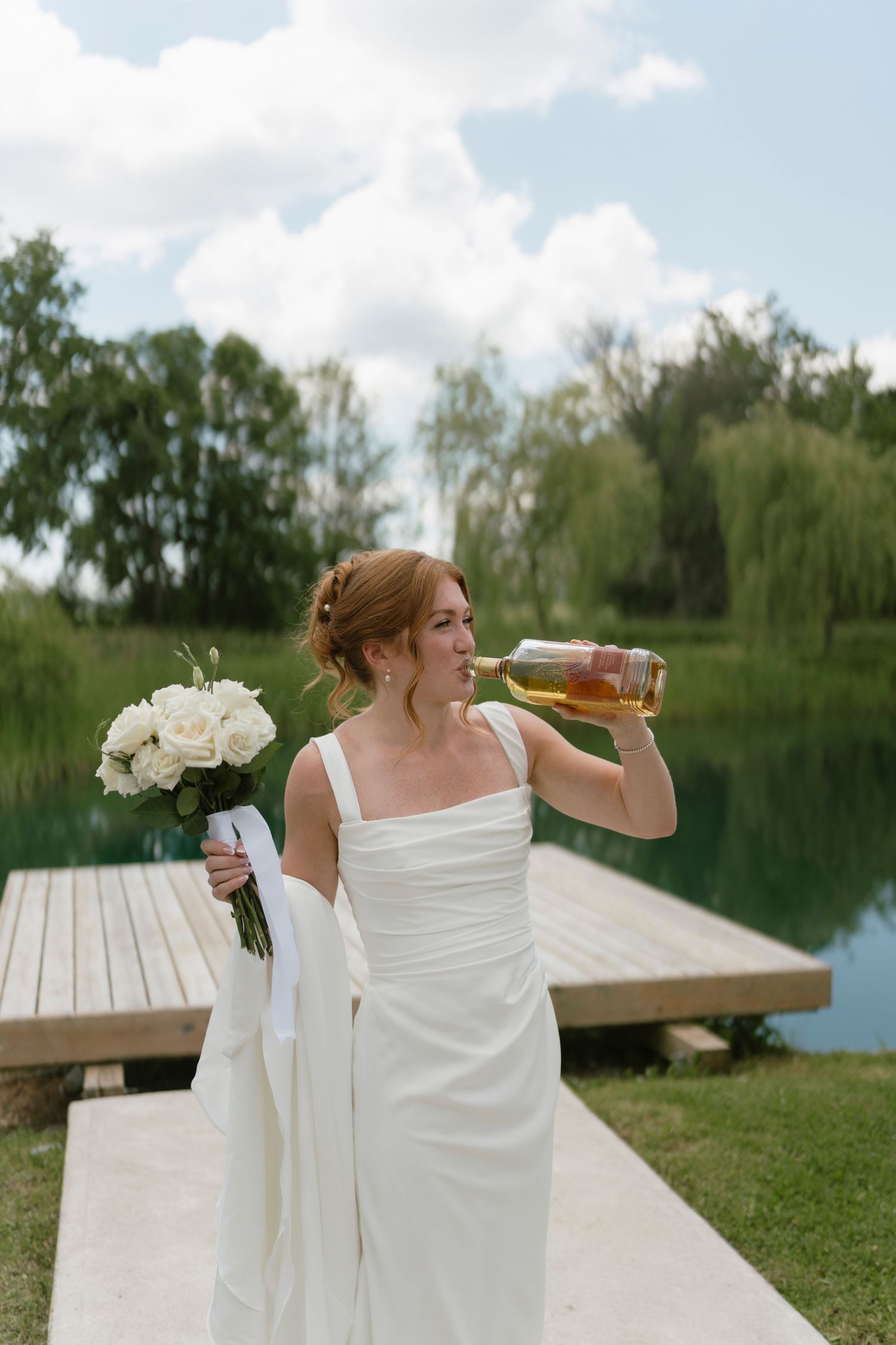 Bride in white gown drinks from bottle while holding white rose bouquet near a serene pond and wooden dock.