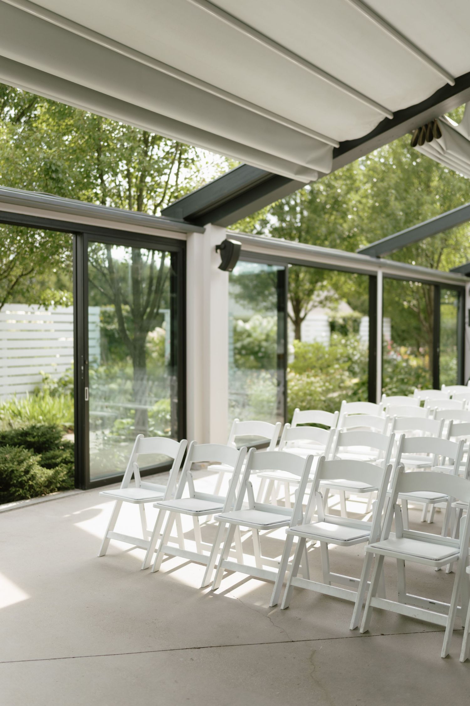 White folding chairs arranged in rows inside a modern glass-walled pavilion surrounded by lush green trees.