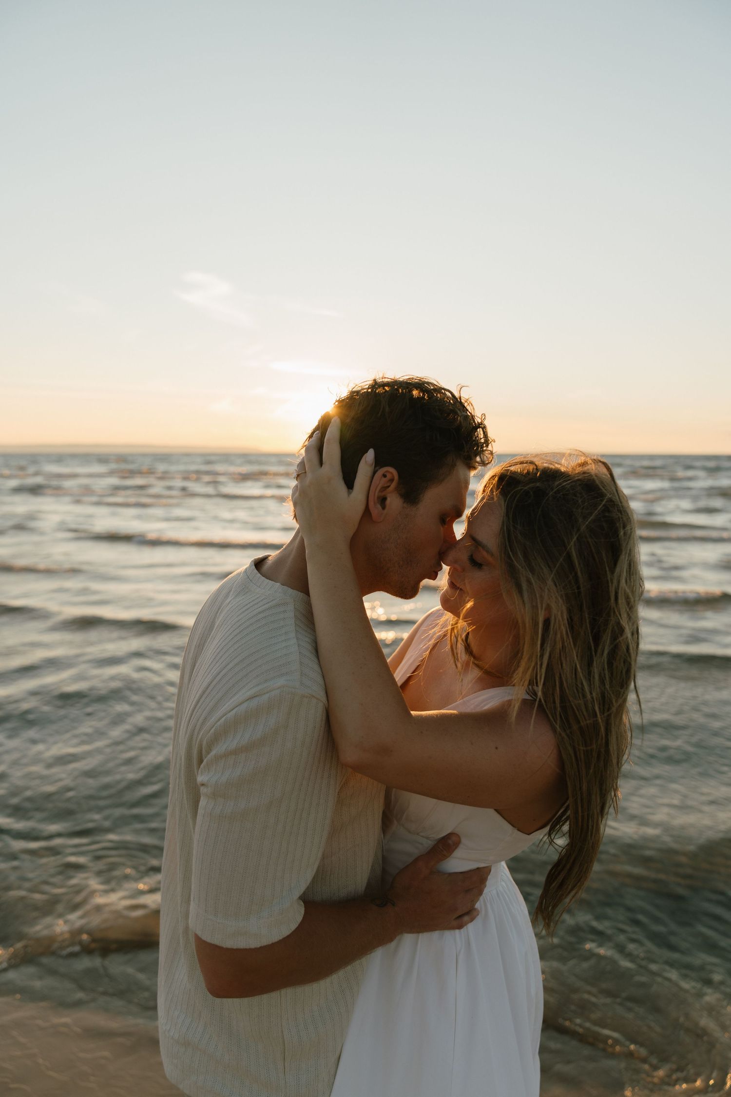 A couple in white clothing shares an intimate moment on the beach at sunset.