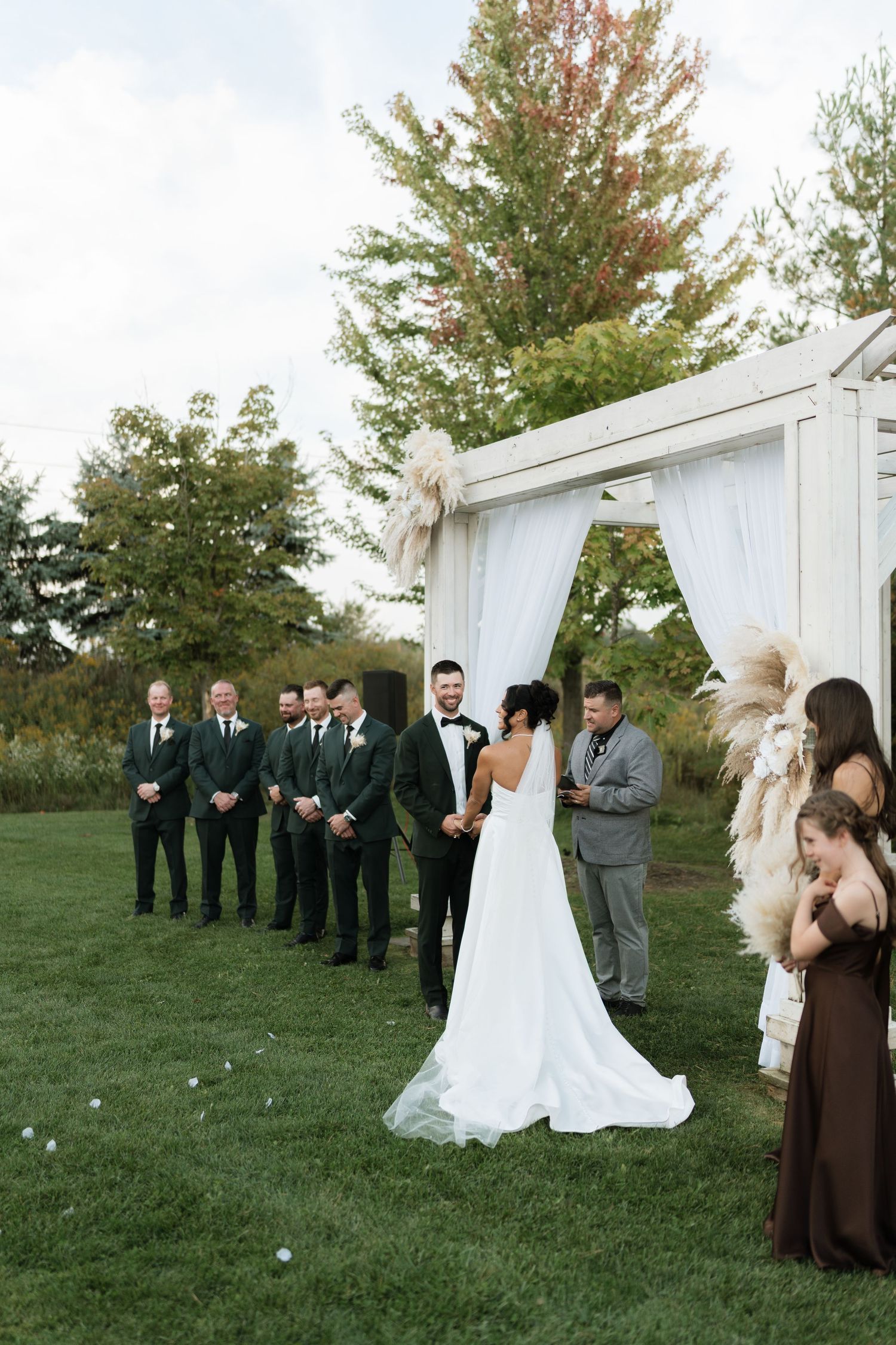 A wedding ceremony setup outdoors with a white arch and wedding party in formal attire.