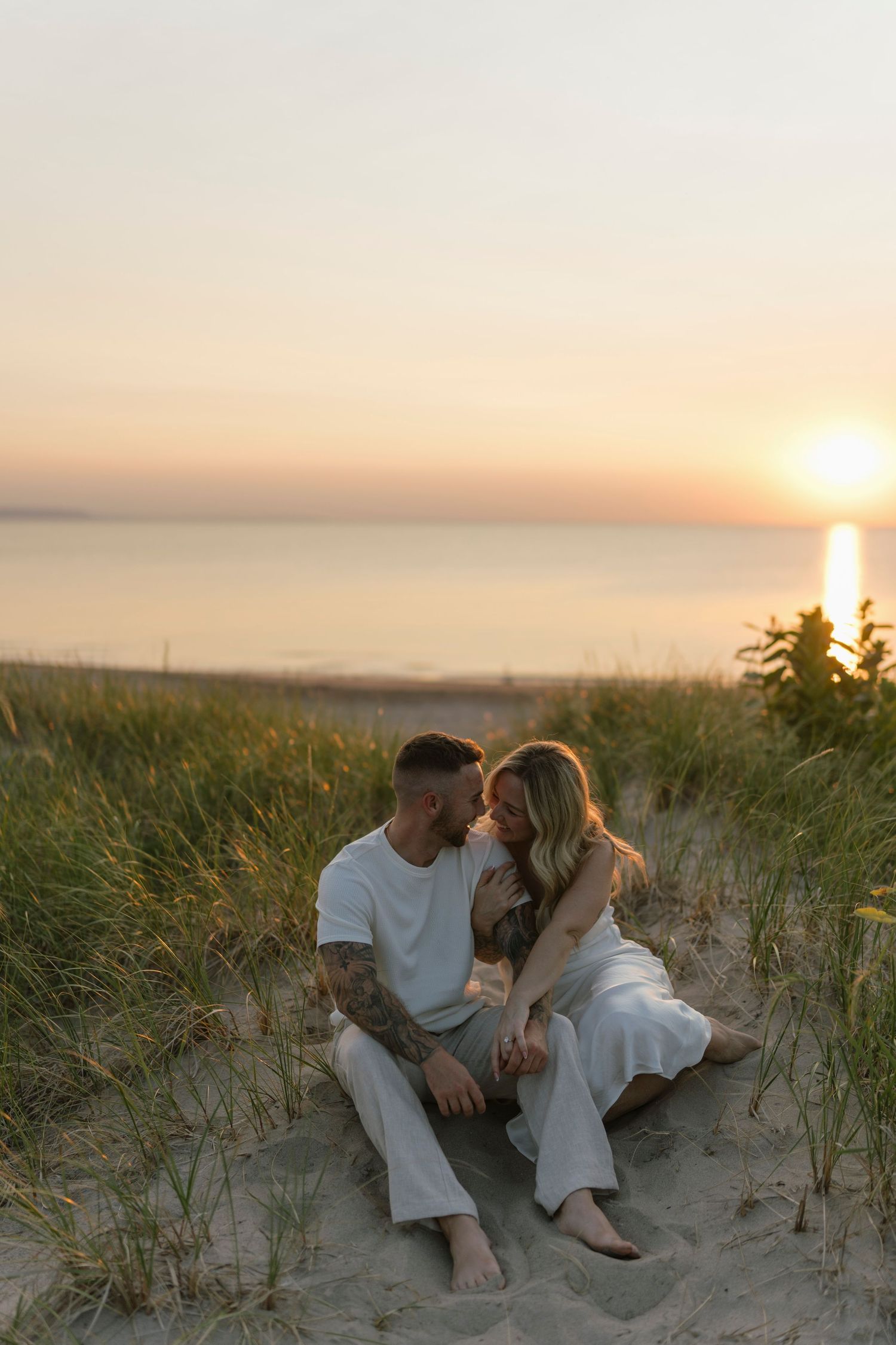 Couple sitting together on sandy beach dunes at golden sunset, sharing a romantic moment with ocean in the background.