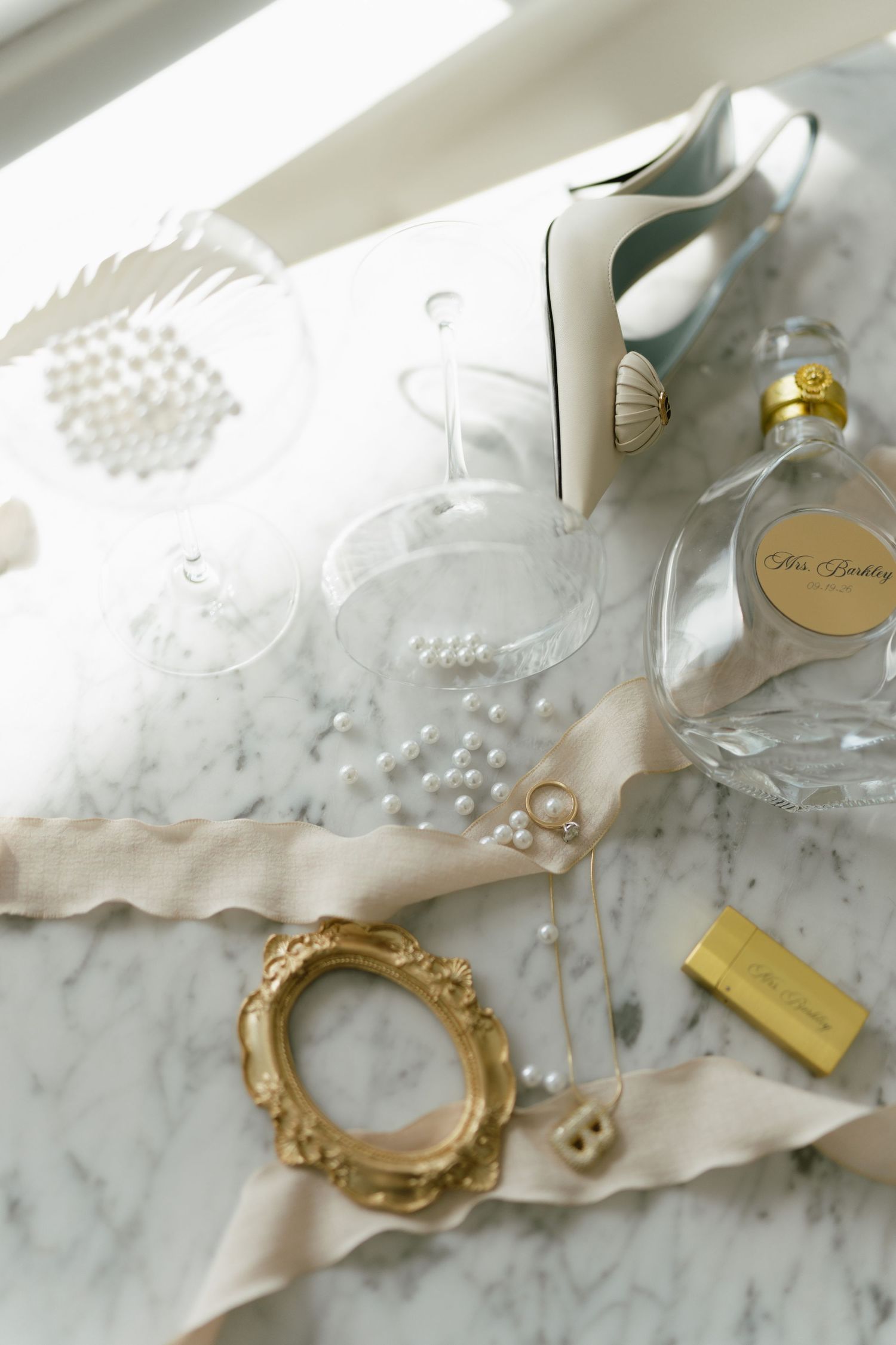 Elegant bridal flat lay with pearl jewelry, champagne flutes, perfume bottle, gold frame, and white heels on marble.