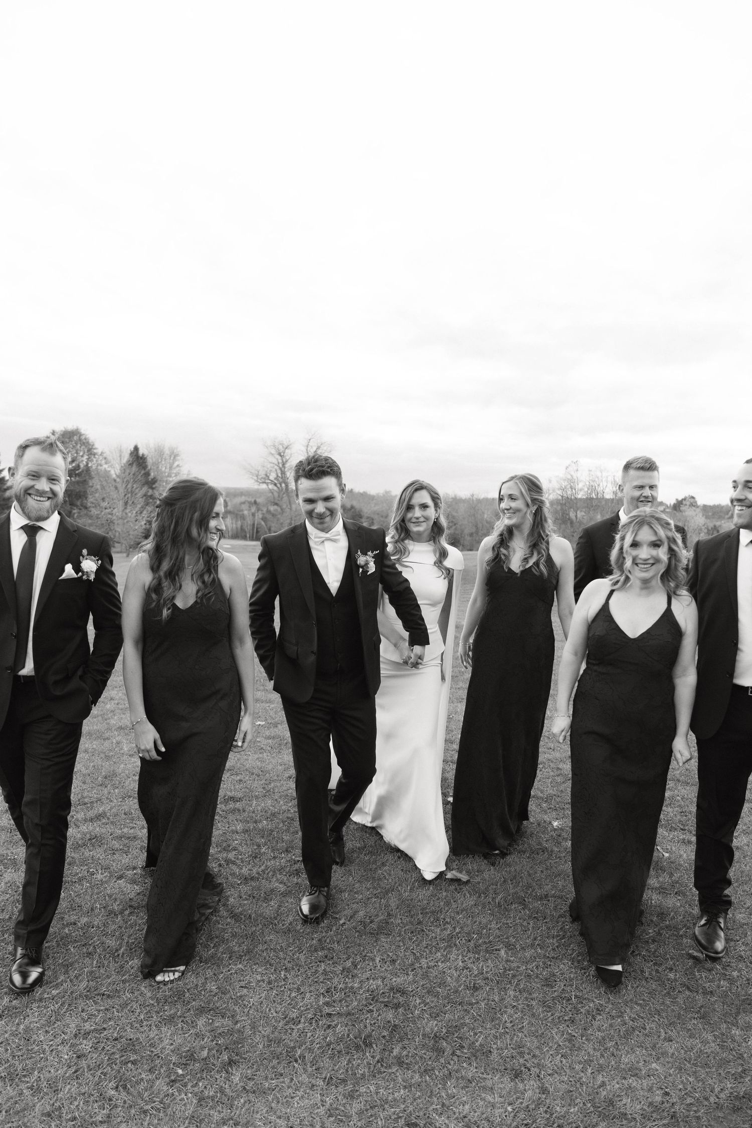 Wedding party walks and laughs together outdoors wearing formal black attire.