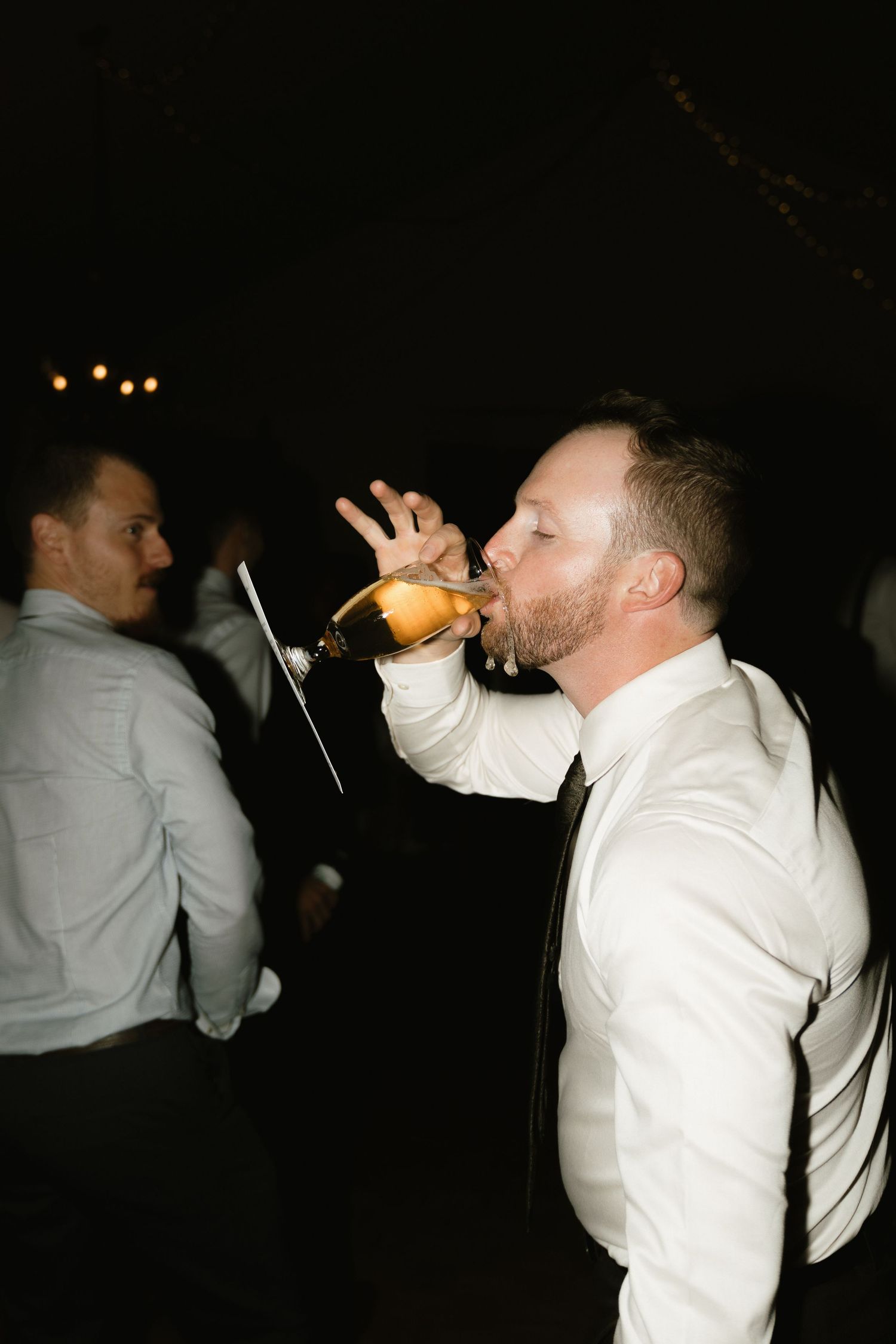 Wedding guest in white shirt drinking from bottle during nighttime reception.