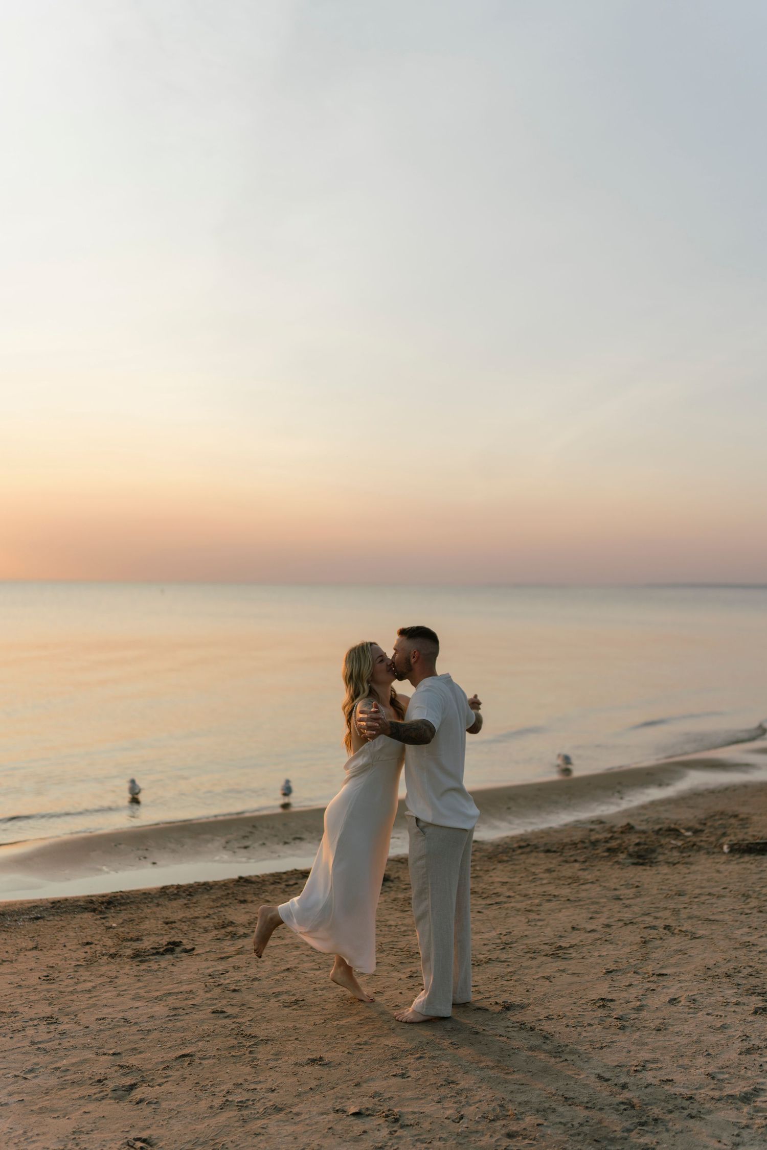 Couple kissing on a sandy beach at sunset, woman in white dress lifted by man in white outfit with ocean backdrop.