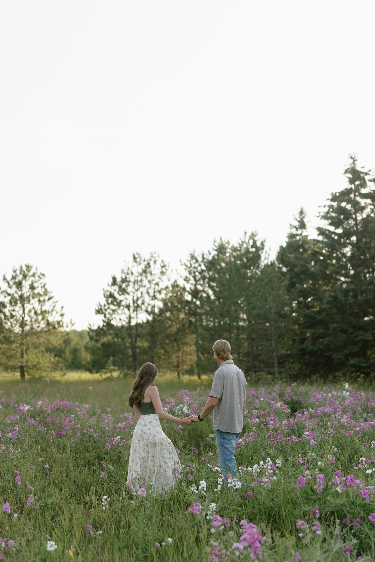 Couple holding hands in a field of pink wildflowers with evergreen trees in the background at sunset.