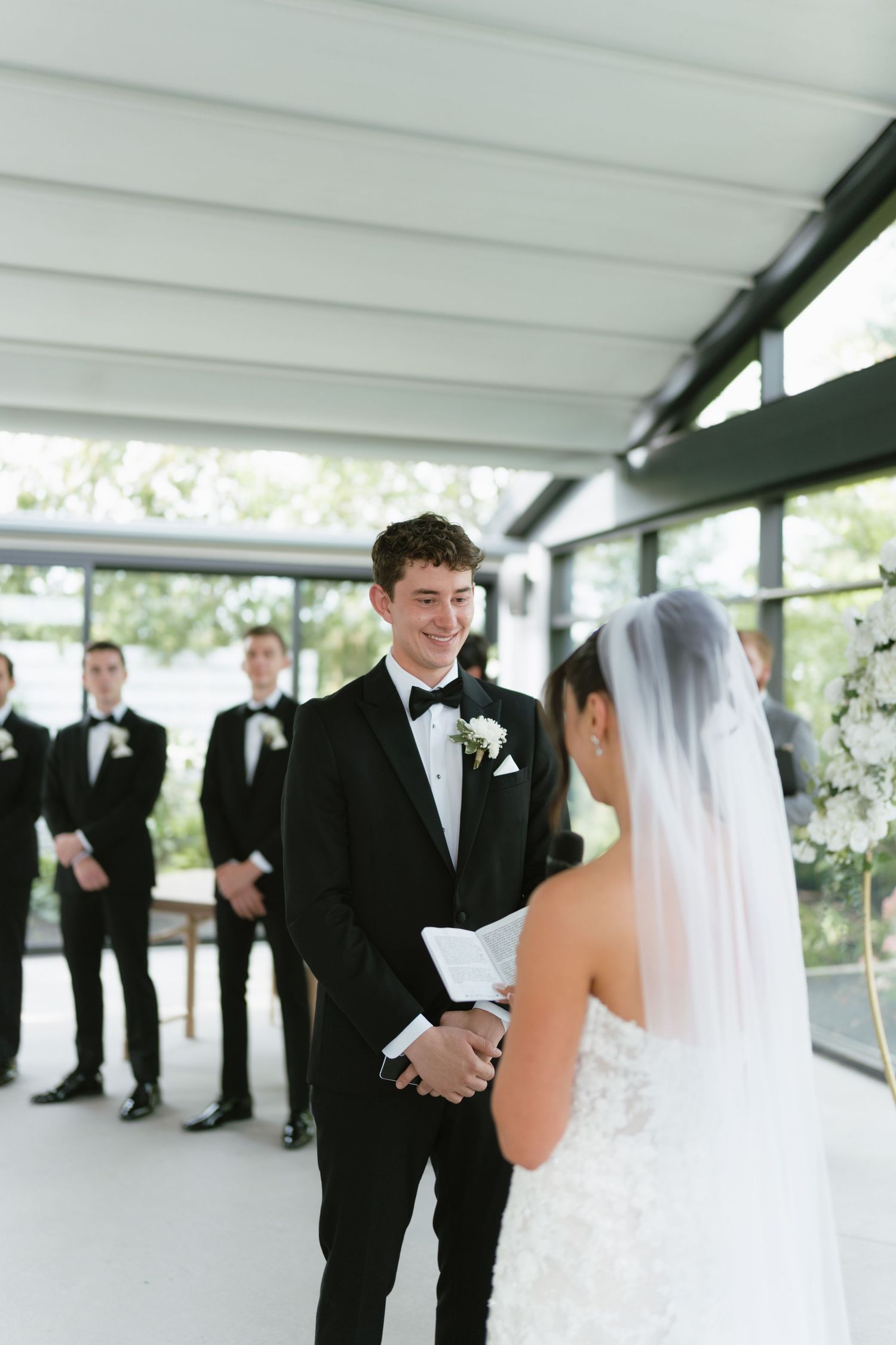 Groom in black tuxedo smiling at bride in white gown and veil during indoor wedding ceremony with groomsmen.